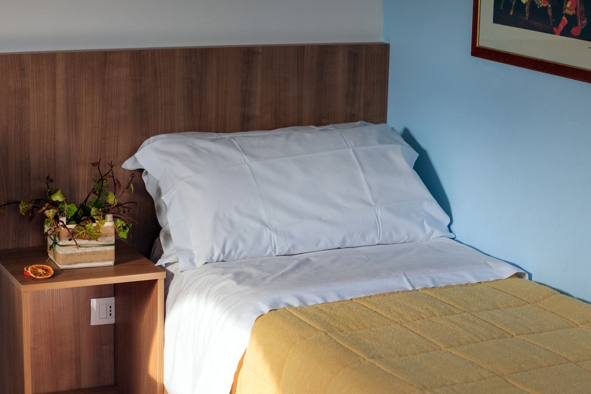 Letto singolo con lenzuola bianche, coperta gialla e testiera in legno, accanto a un comodino con fiori e una lampada.