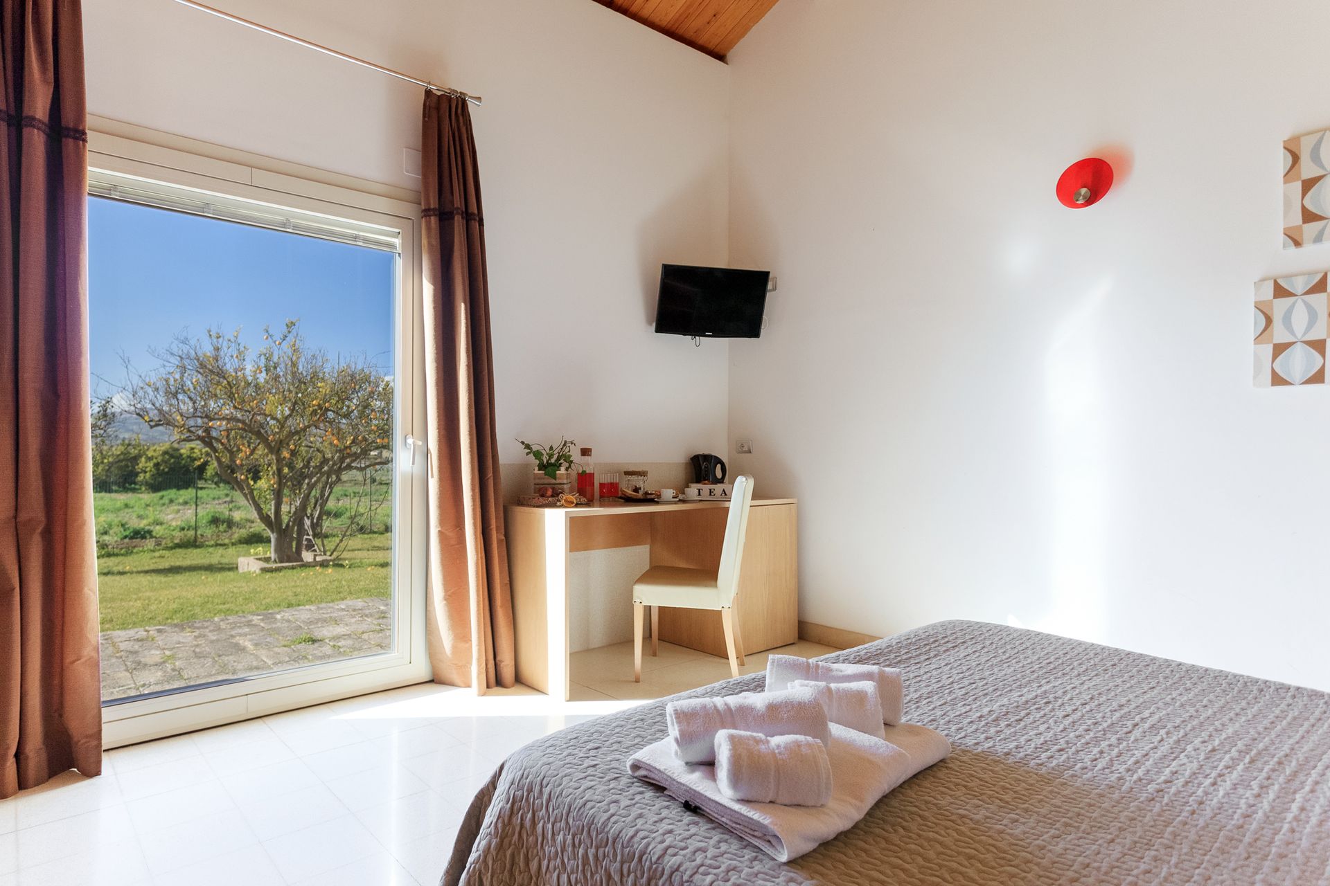 Luminosa camera da letto con letto, scrivania, TV a parete e ampia finestra con vista su un cortile verde.