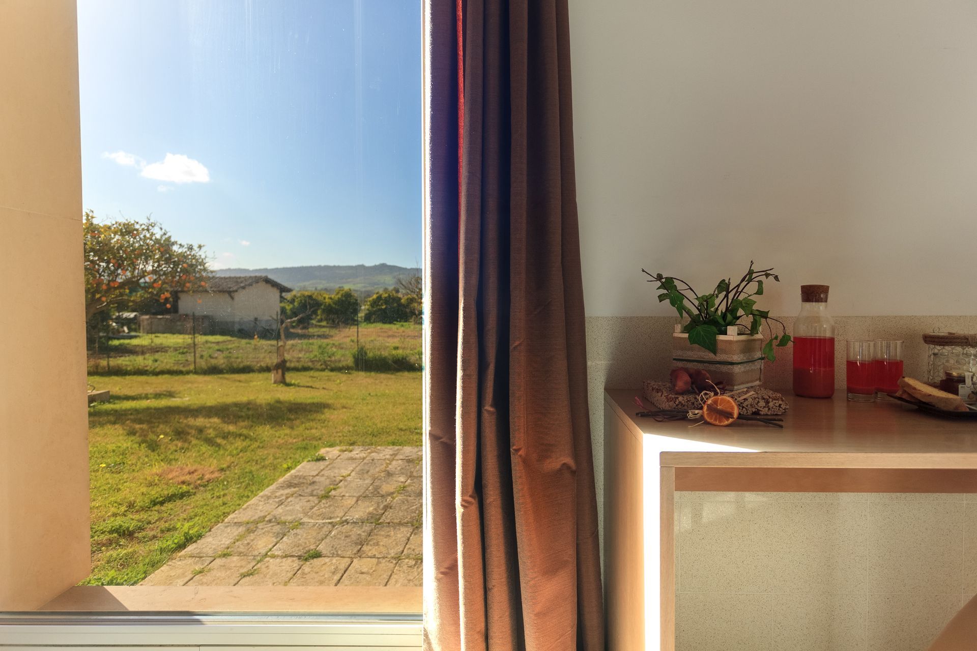 Vista attraverso una finestra aperta su un giardino soleggiato, con tende e un tavolino.