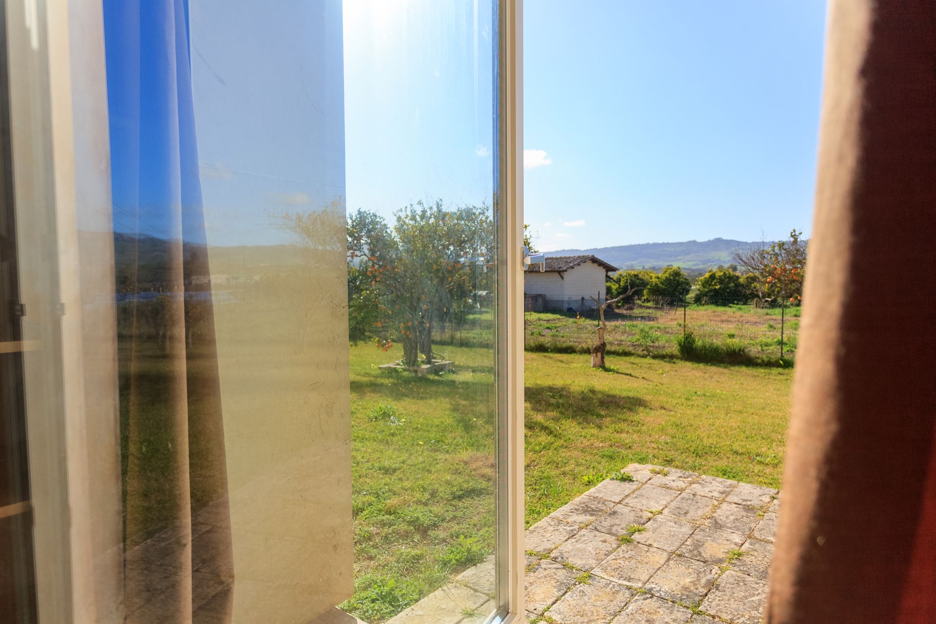 Vista attraverso una finestra di un cortile soleggiato con prato, alberi, un patio e colline in lontananza.