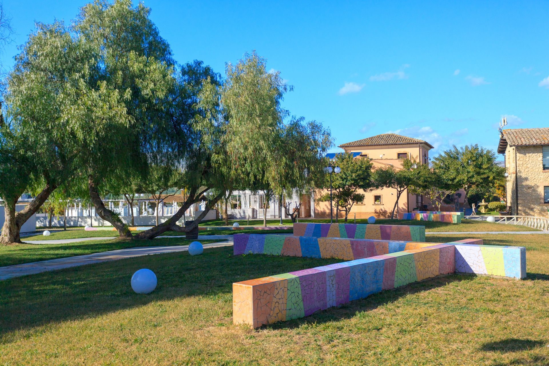 Sedute colorate in un parco soleggiato con alberi, prato ed edifici sullo sfondo.
