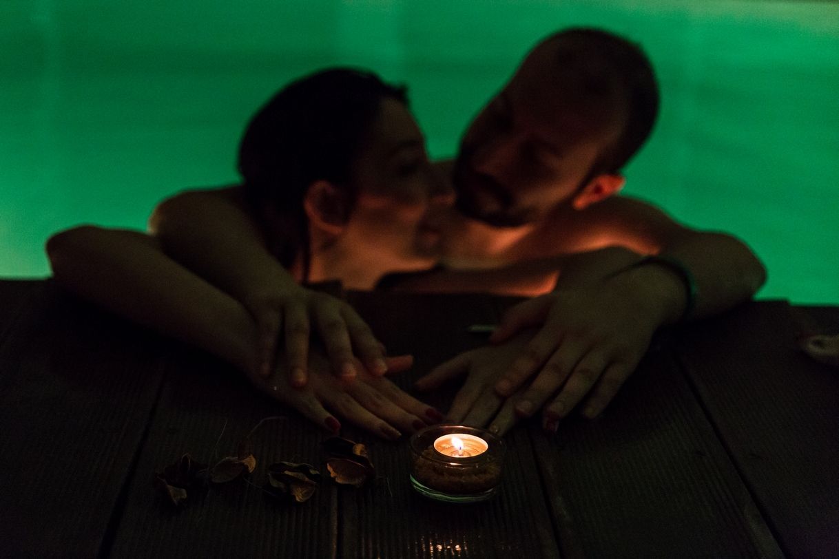 Due persone si baciano a lume di candela su un tavolo cosparso di petali e con uno sfondo illuminato di verde.