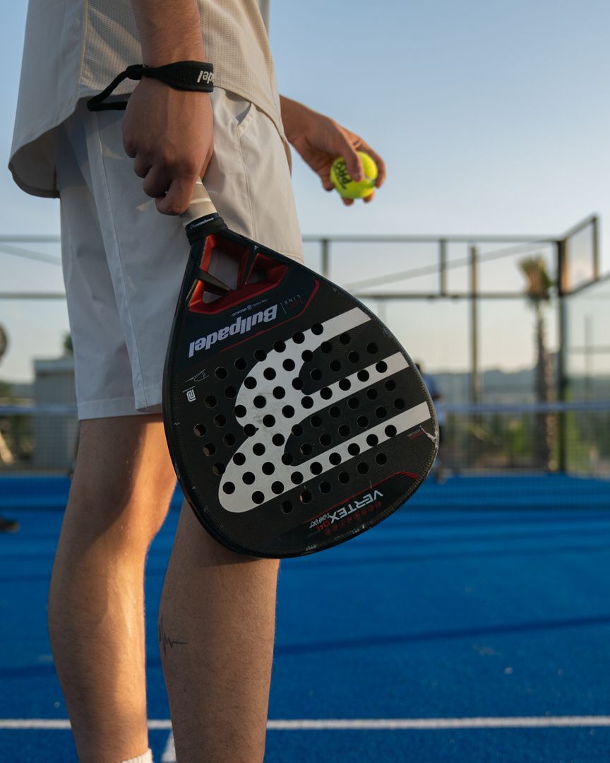 Una persona tiene in mano una racchetta da pickleball nera e una palla gialla su un campo blu all'aperto al tramonto.