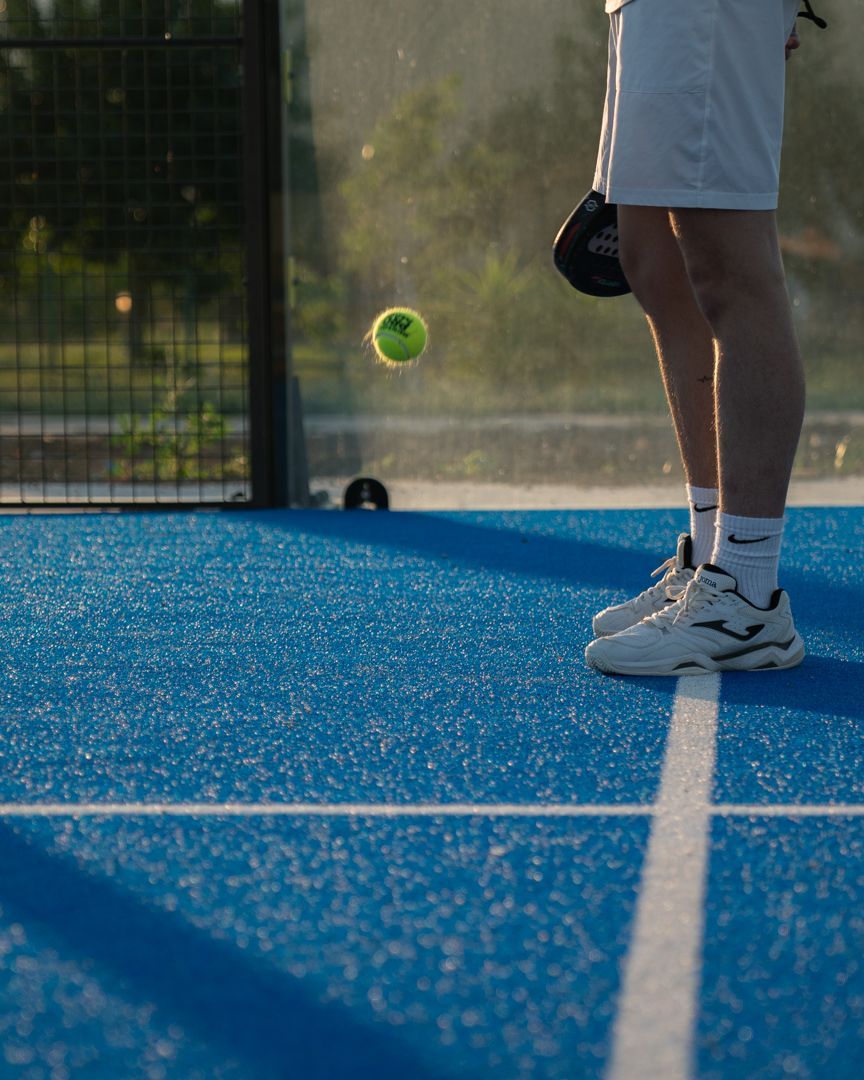 Gambe e racchetta di un tennista su un campo blu mentre una palla gialla rimbalza vicino alla rete.