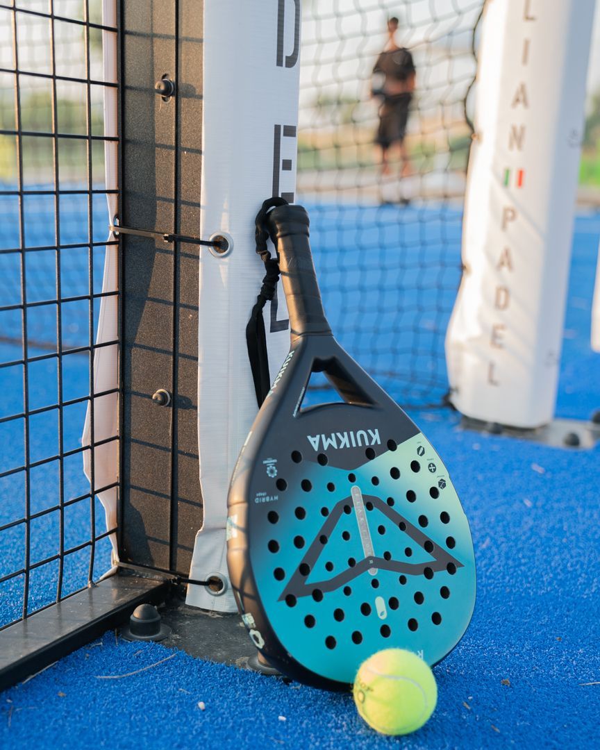 Una racchetta da padel appoggiata a un palo della rete su un campo blu, con una pallina da tennis nelle vicinanze.