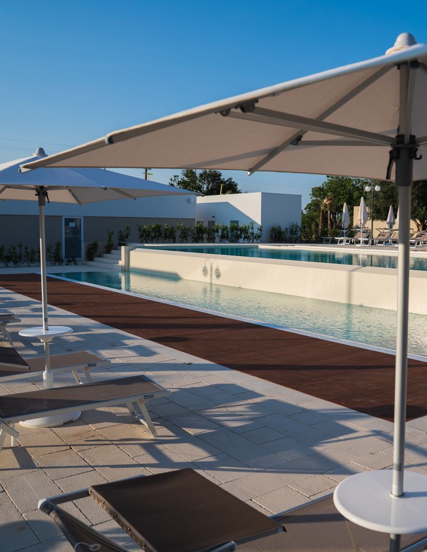 Lettini a bordo piscina sotto grandi ombrelloni beige, accanto a una piscina a sfioro turchese sotto un cielo azzurro.