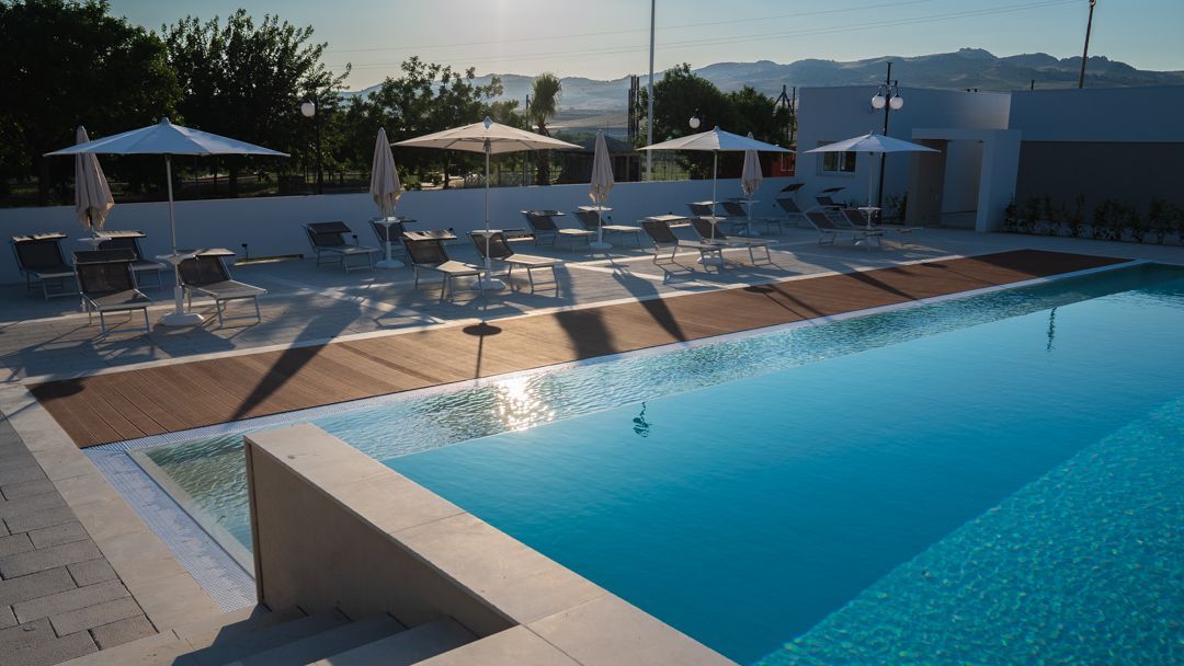 Piscina all'aperto del resort con lettini, ombrelloni e ombrelloni che si riflettono nell'acqua blu al tramonto.