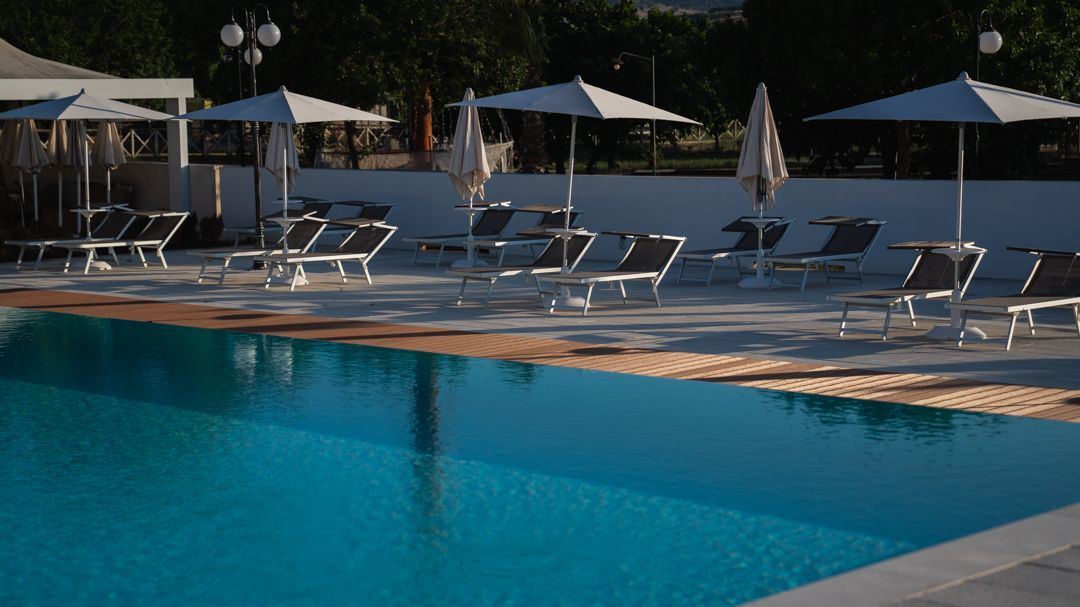 Piscina del resort con palma, lettini e terrazza in legno sotto un cielo azzurro limpido.