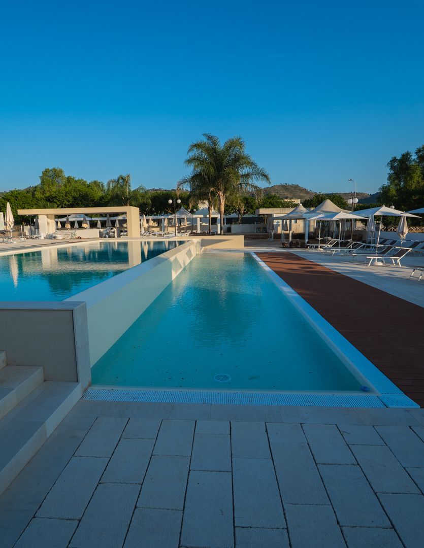 Piscina all'aperto del resort con lettini, ombrelloni e ombrelloni che si riflettono nell'acqua blu al tramonto.