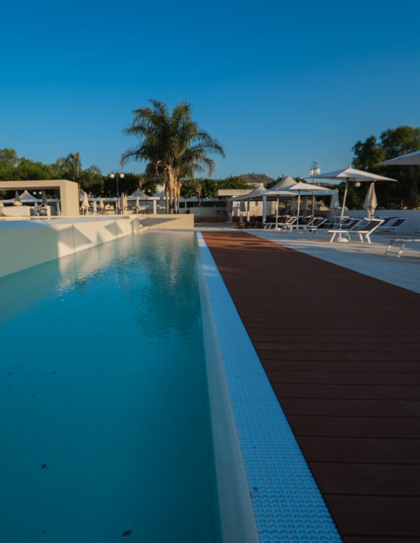 Piscina del resort con palma, lettini e terrazza in legno sotto un cielo azzurro limpido.
