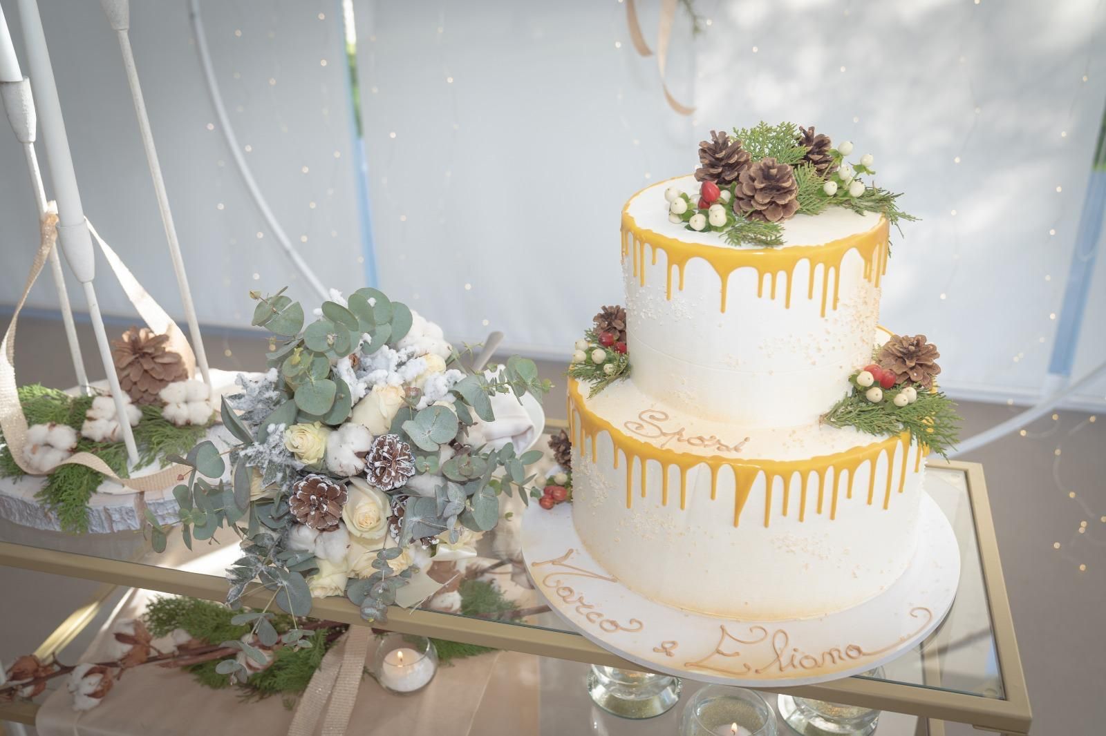 Torta nuziale bianca a due piani con glassa dorata a goccia e decorazioni floreali su un tavolo di vetro.