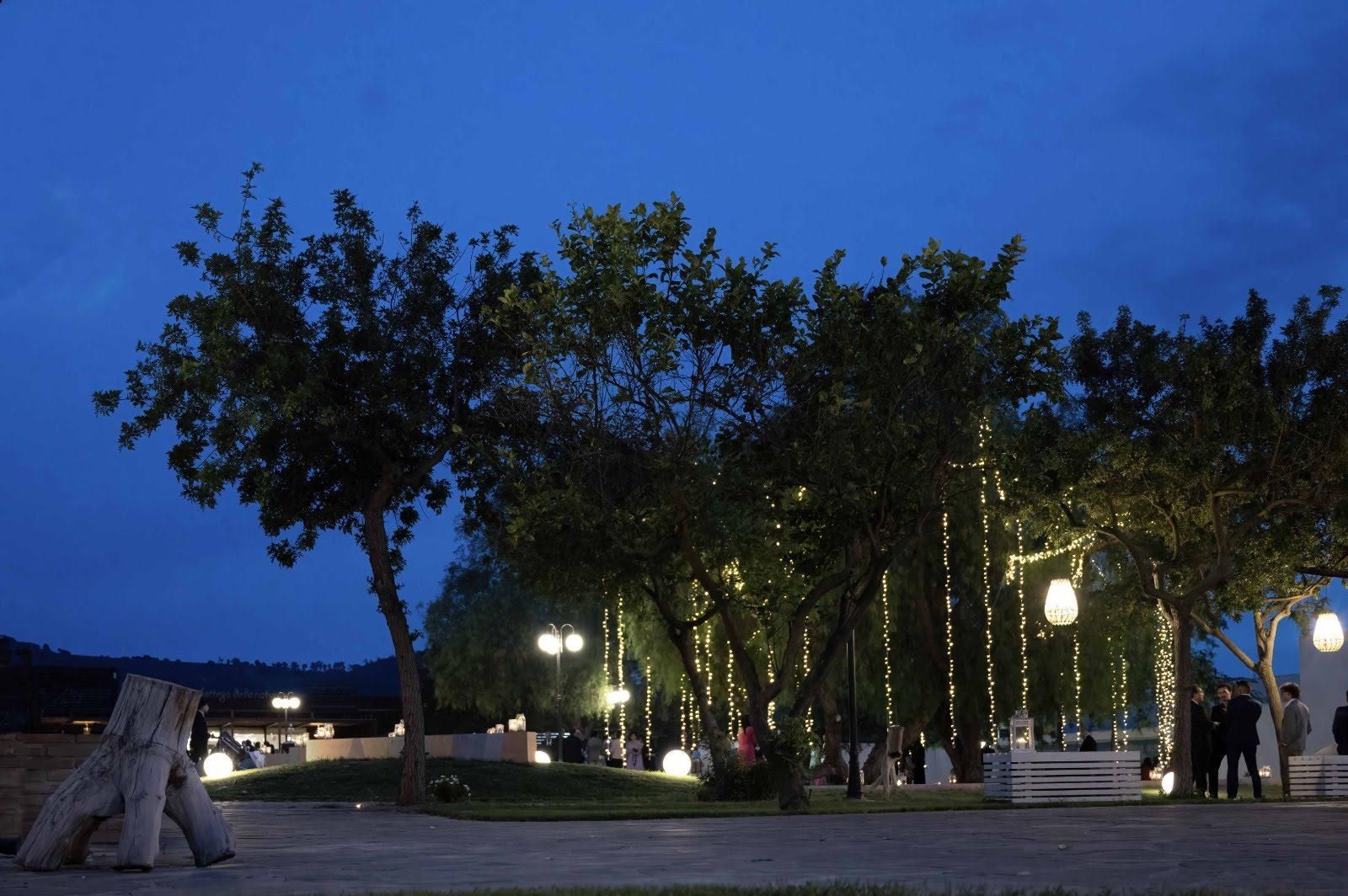 Parco crepuscolare con alberi, sentiero illuminato e un piccolo padiglione bianco sotto il cielo azzurro della sera.