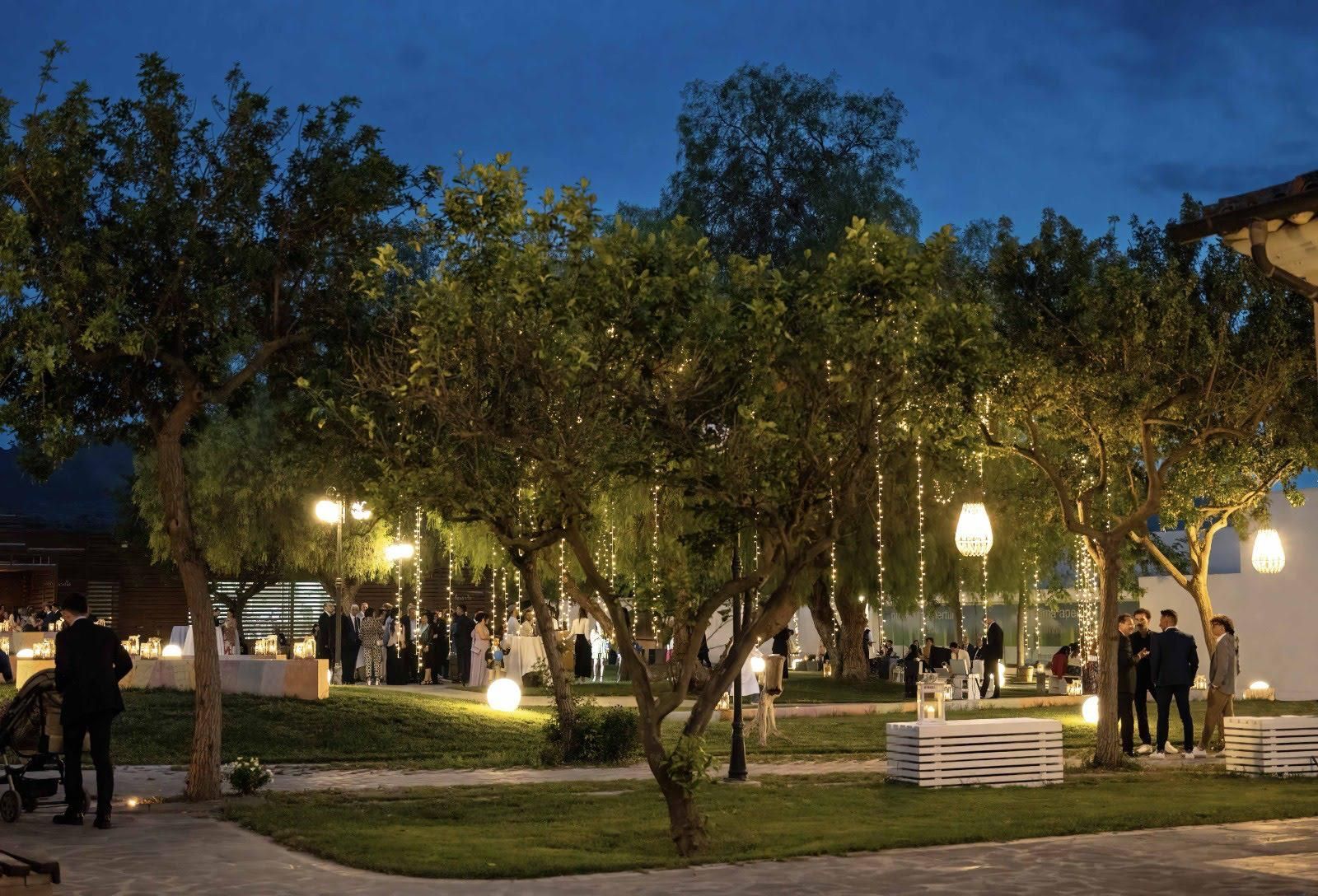 Parco illuminato al crepuscolo con alberi, panchine e alcune persone che passeggiano vicino ai lampioni.