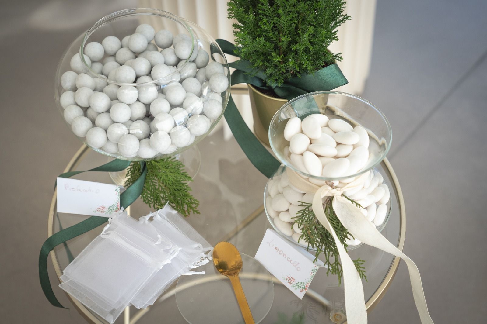 Ciotole di vetro decorative con caramelle bianche e nastri su un tavolo, con piccole piante verdi ed etichette regalo.