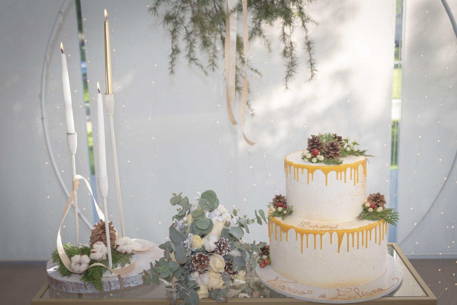 Torta nuziale bianca con decorazioni a goccia dorata accanto a un centrotavola floreale con candele su un tavolo decorato.