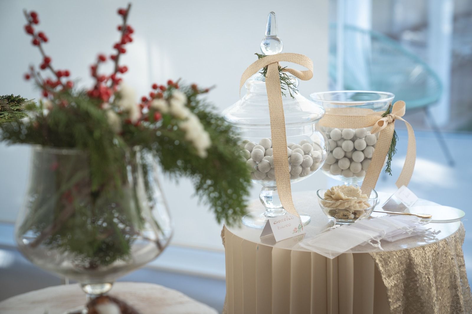 Allestimento per tavolo nuziale color crema con vaso di vetro, nastro bianco e ciotola con perle vicino a una finestra.