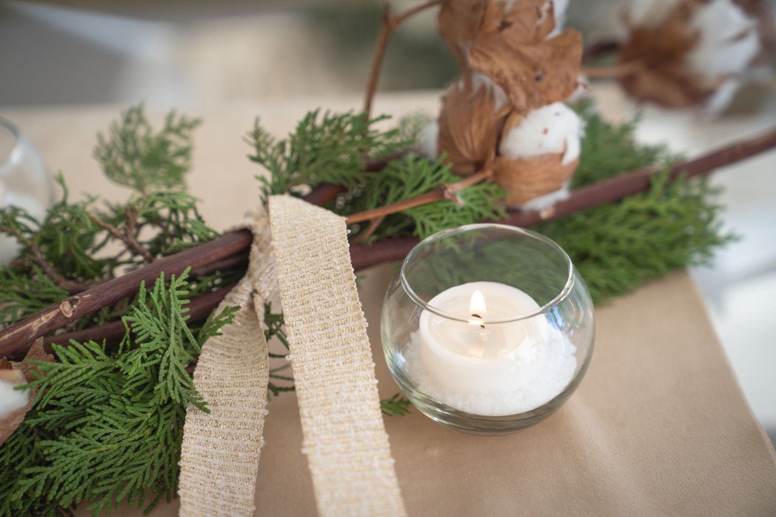 Candele e verde disposti su un runner da tavola beige con rametti di legno e un portacandele in vetro.