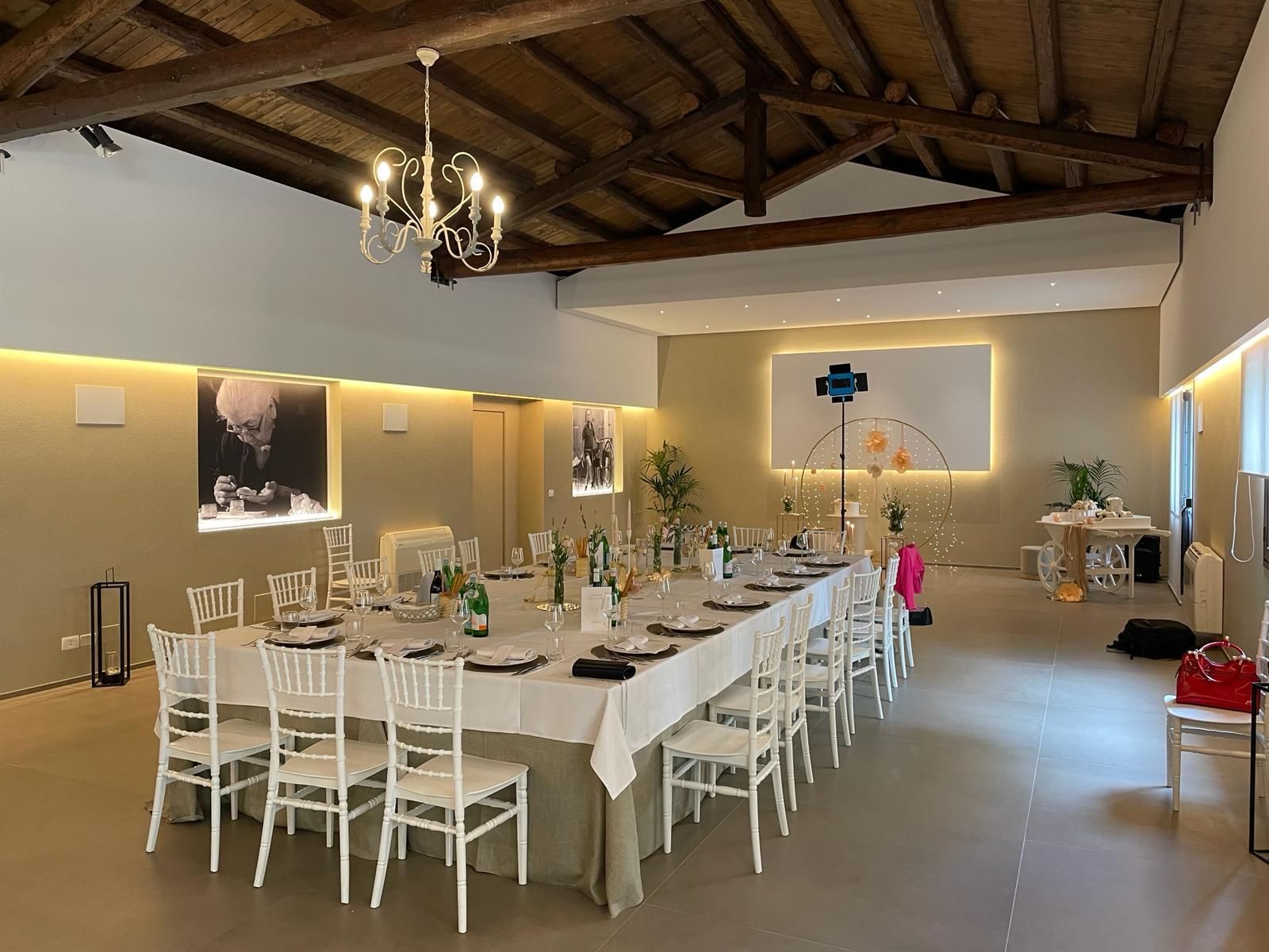 Elegante sala da pranzo con un lungo tavolo apparecchiato, sedie bianche, lampadari e soffitto rustico con travi in ​​legno.