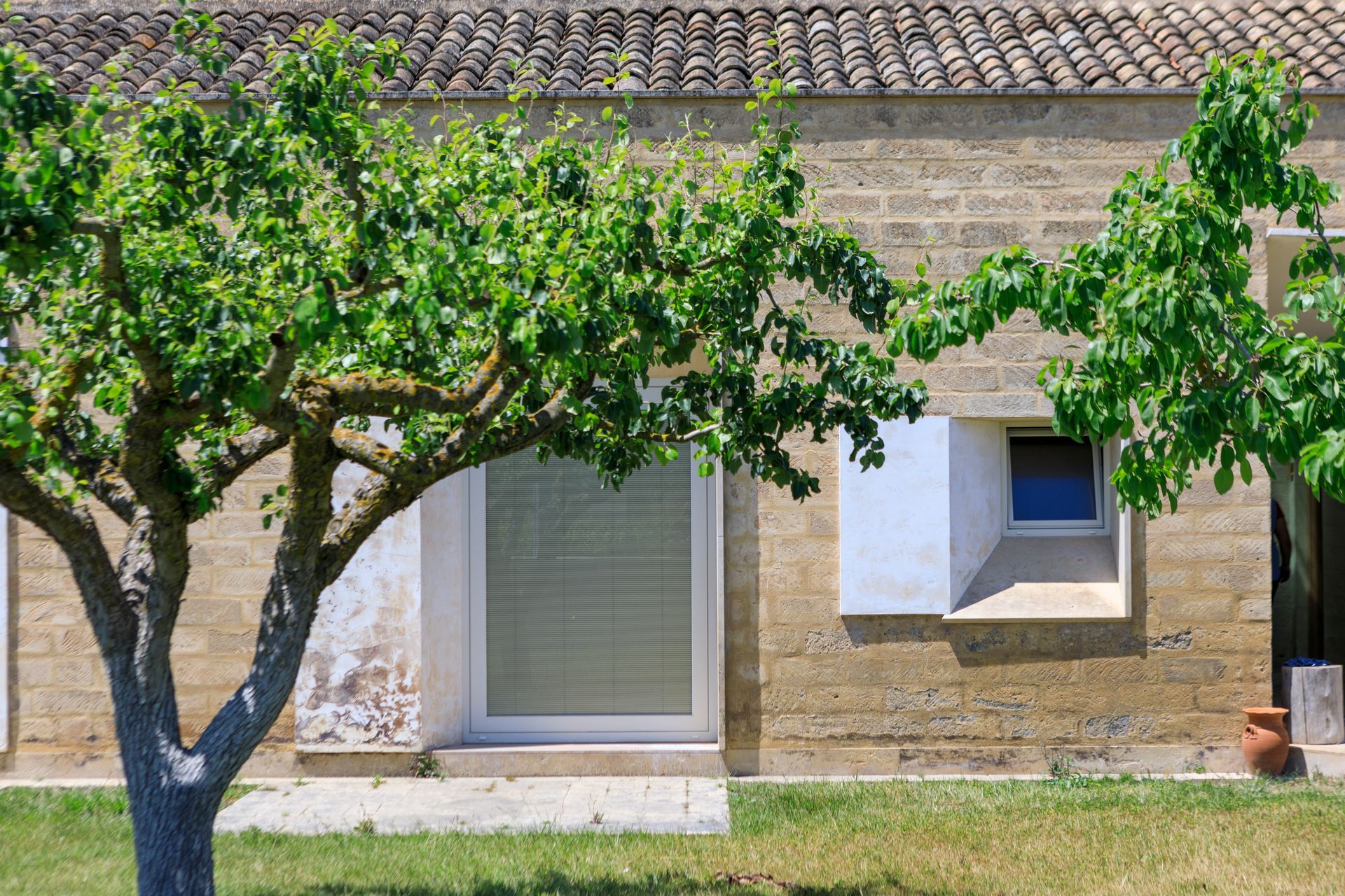 Casa in pietra soleggiata con due alberi frondosi davanti e una piccola finestra con persiane bianche.