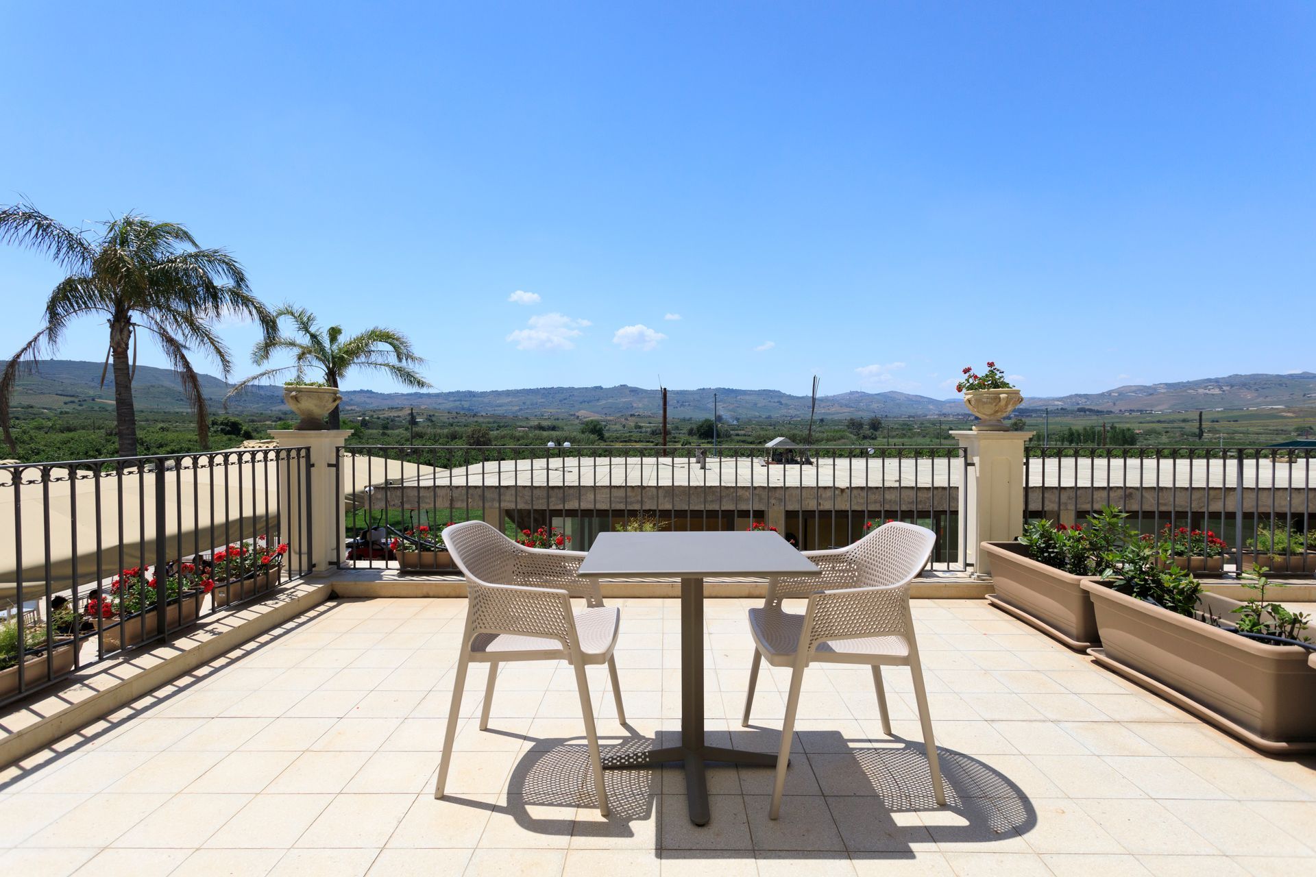Patio con tavolo e due sedie con vista sui giardini e sulle colline in lontananza, sotto un cielo azzurro.