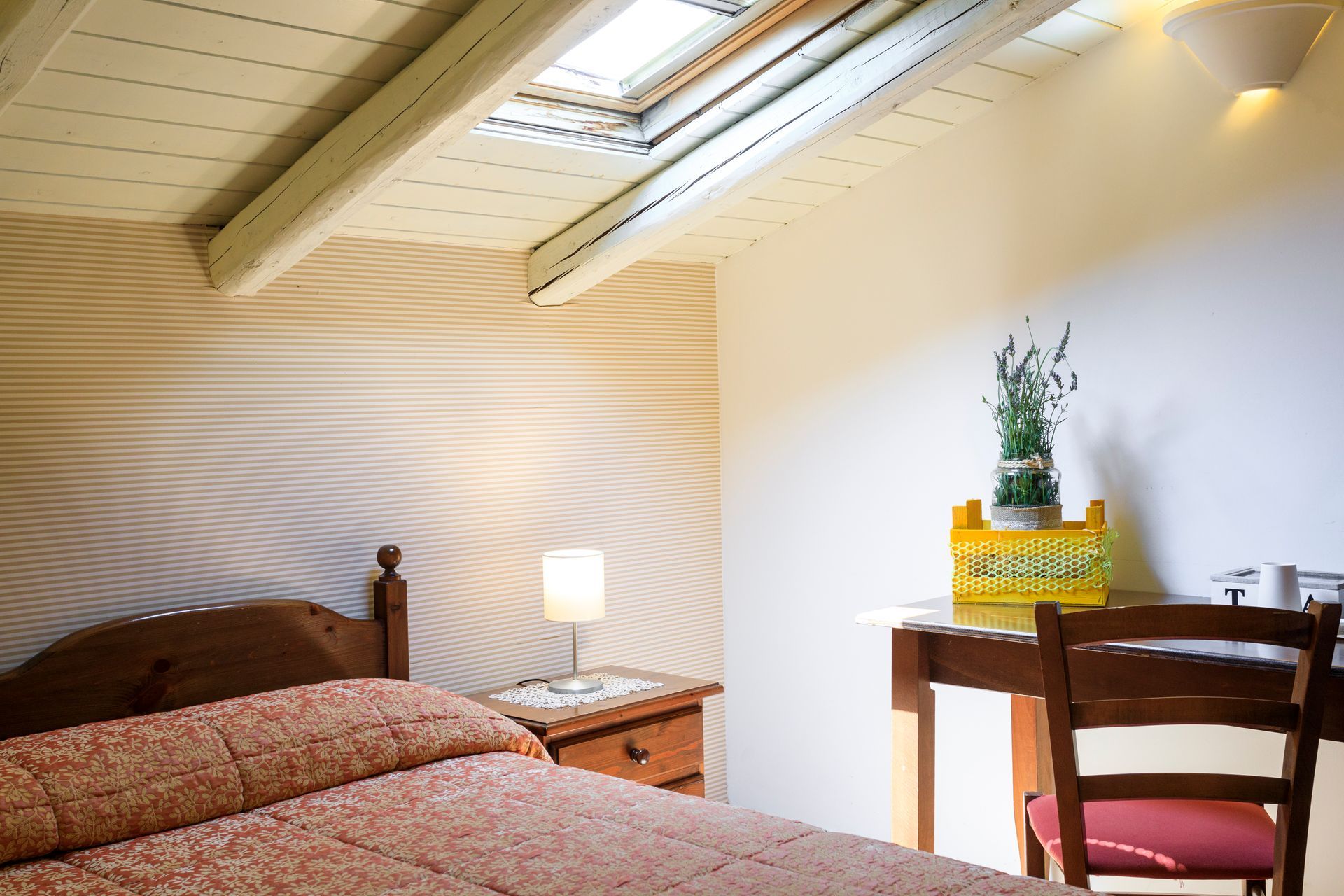 Camera da letto soleggiata con letto, lampada da comodino, lucernari inclinati e una piccola scrivania a muro