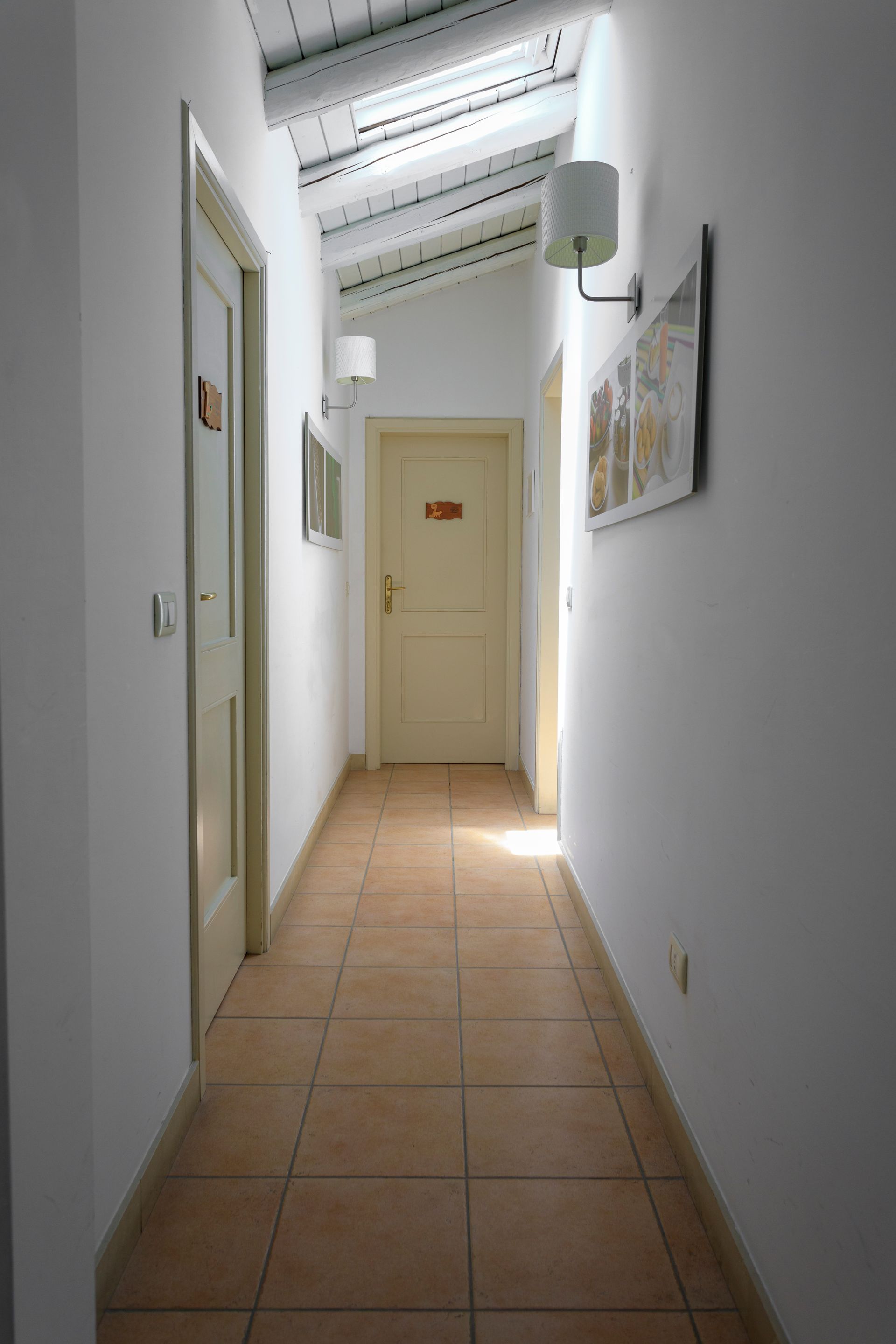 Stretto corridoio bianco con pavimento piastrellato, porte su entrambi i lati e una finestra.