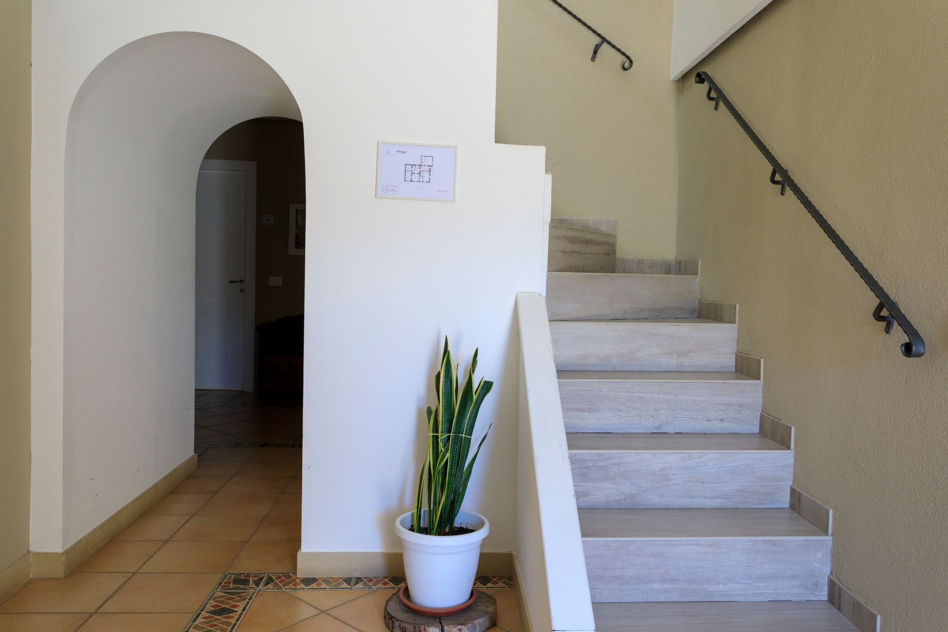 Vano scale interno con pareti beige, pavimento piastrellato, porta ad arco e una pianta in vaso accanto alle scale.