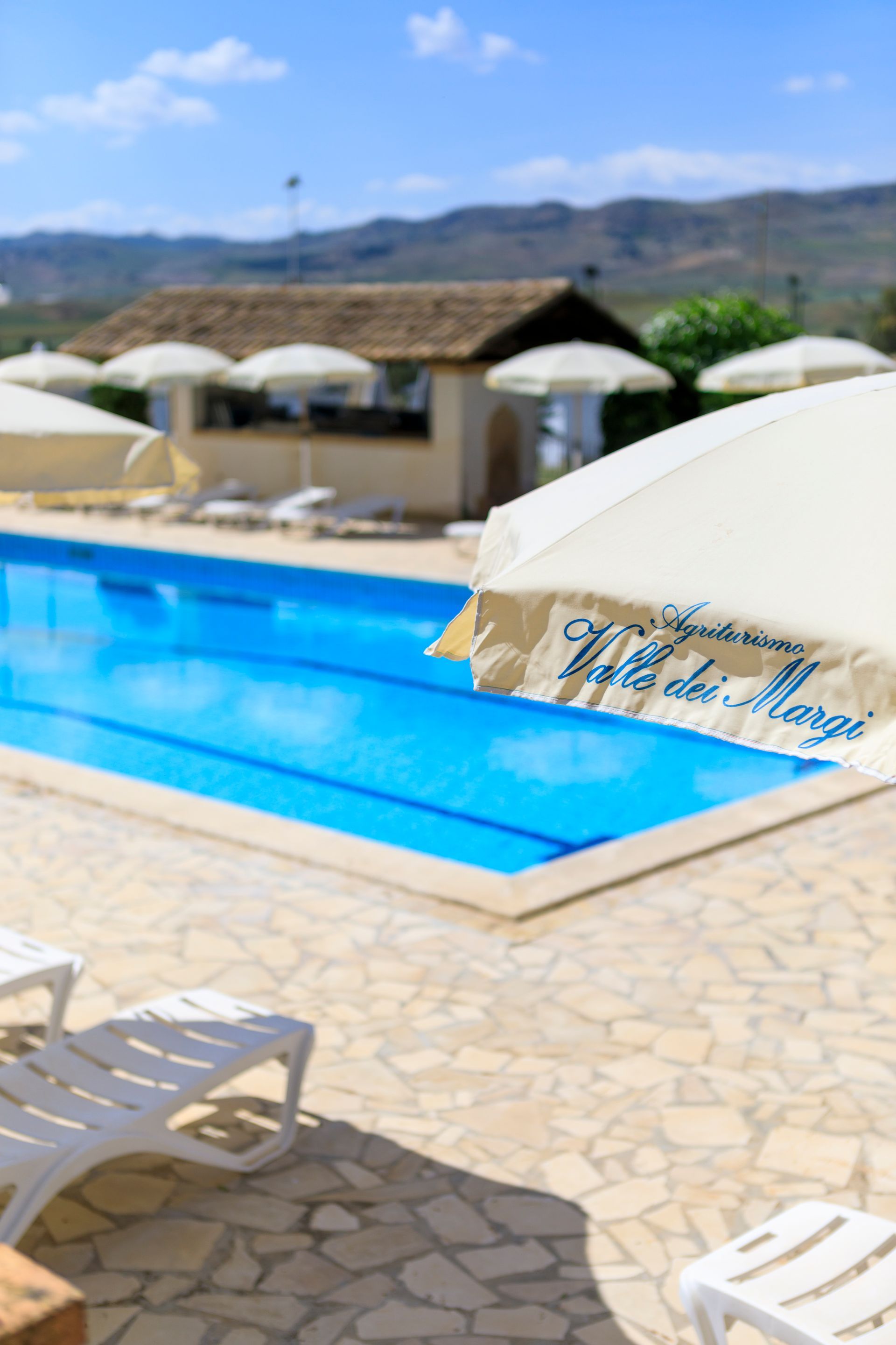 Piscina all'aperto del resort con acqua azzurra, lettini bianchi e ombrelloni sotto un cielo soleggiato.