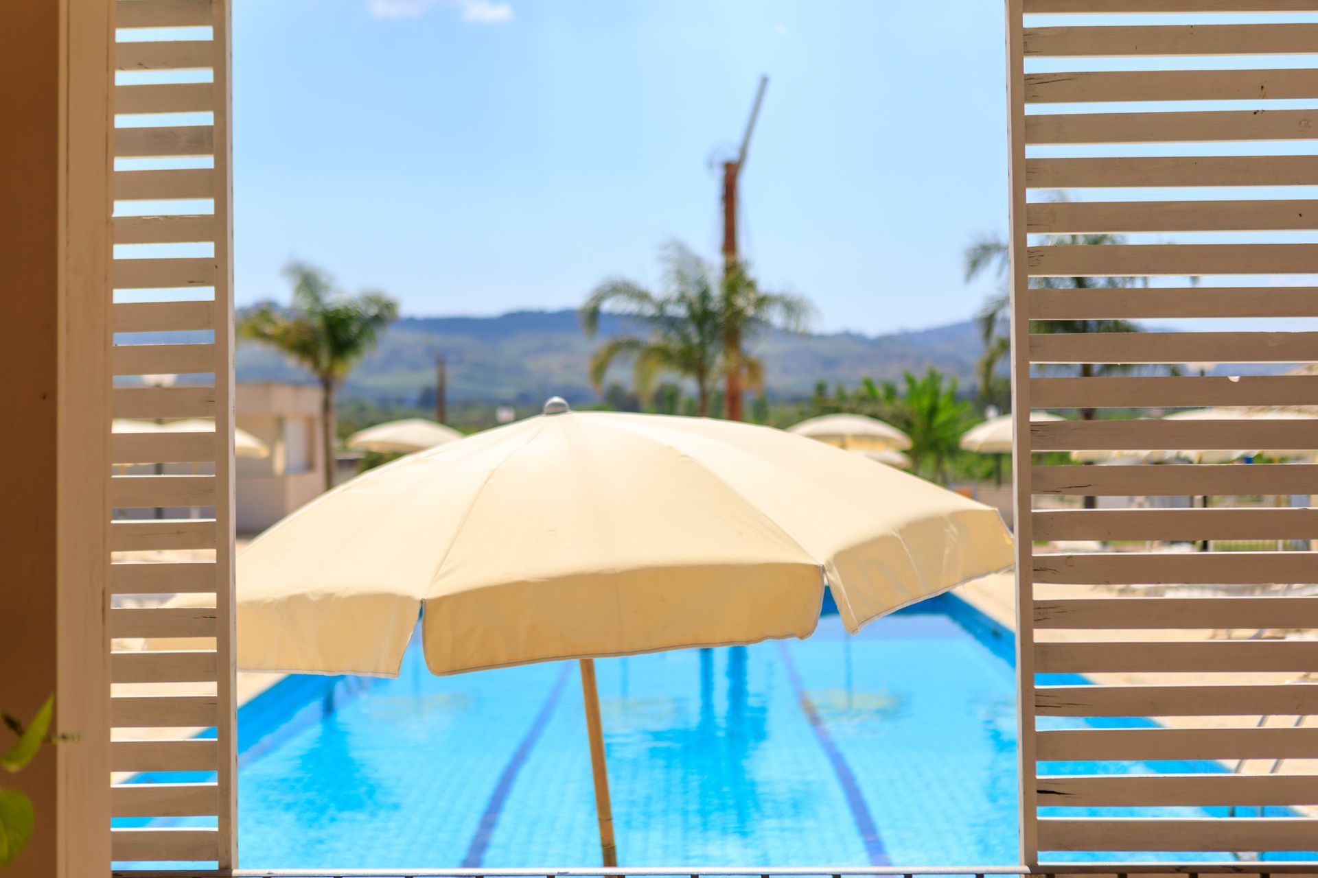 Vista a bordo piscina attraverso le persiane, con un ombrellone beige e acqua blu, palme e oceano in lontananza.