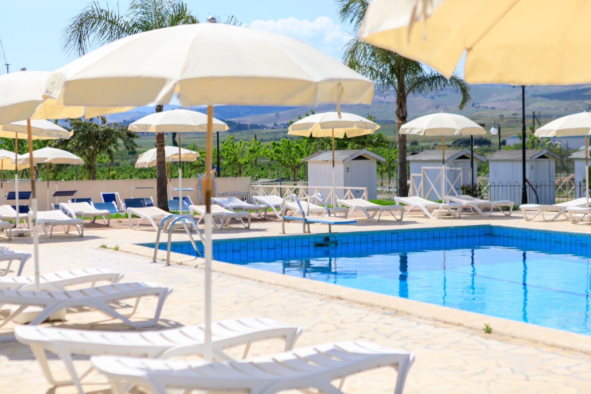 Resort soleggiato a bordo piscina con lettini bianchi, ombrelloni e una piscina blu.
