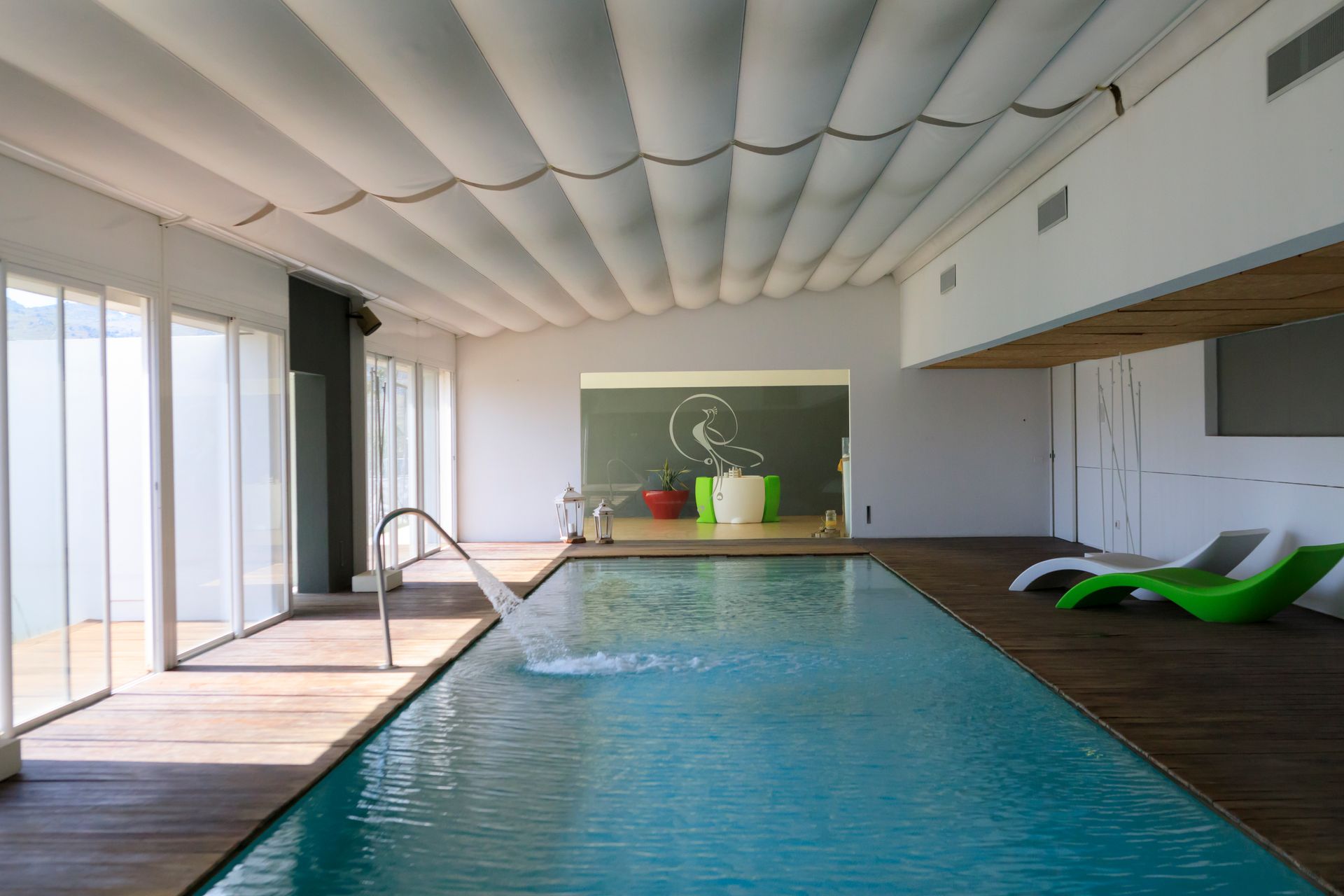 Piscina coperta con acqua turchese, terrazza in legno e lettini prendisole accanto a grandi finestre.