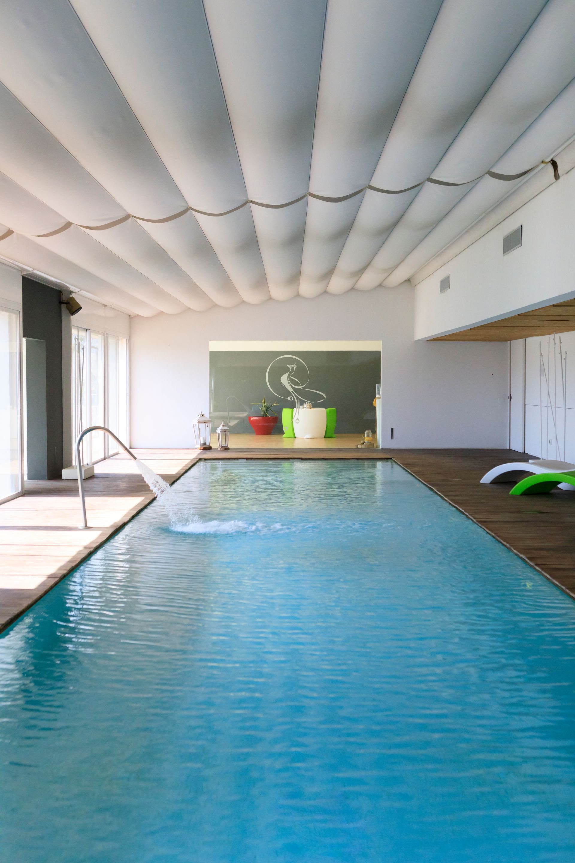 Piscina coperta con acqua blu, pannelli bianchi sul soffitto e una piccola area salotto all'estremità.
