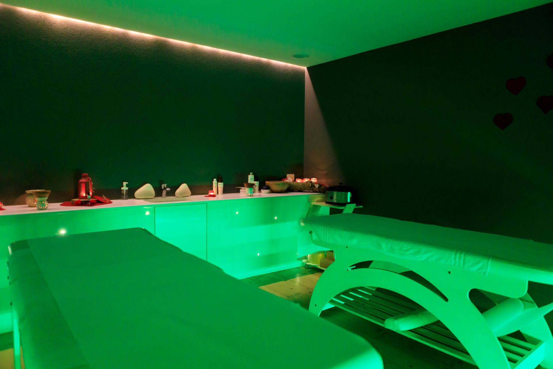 Stanza illuminata di verde con scaffali pieni di piccoli oggetti, parete nera e un letto o lettino da massaggio.