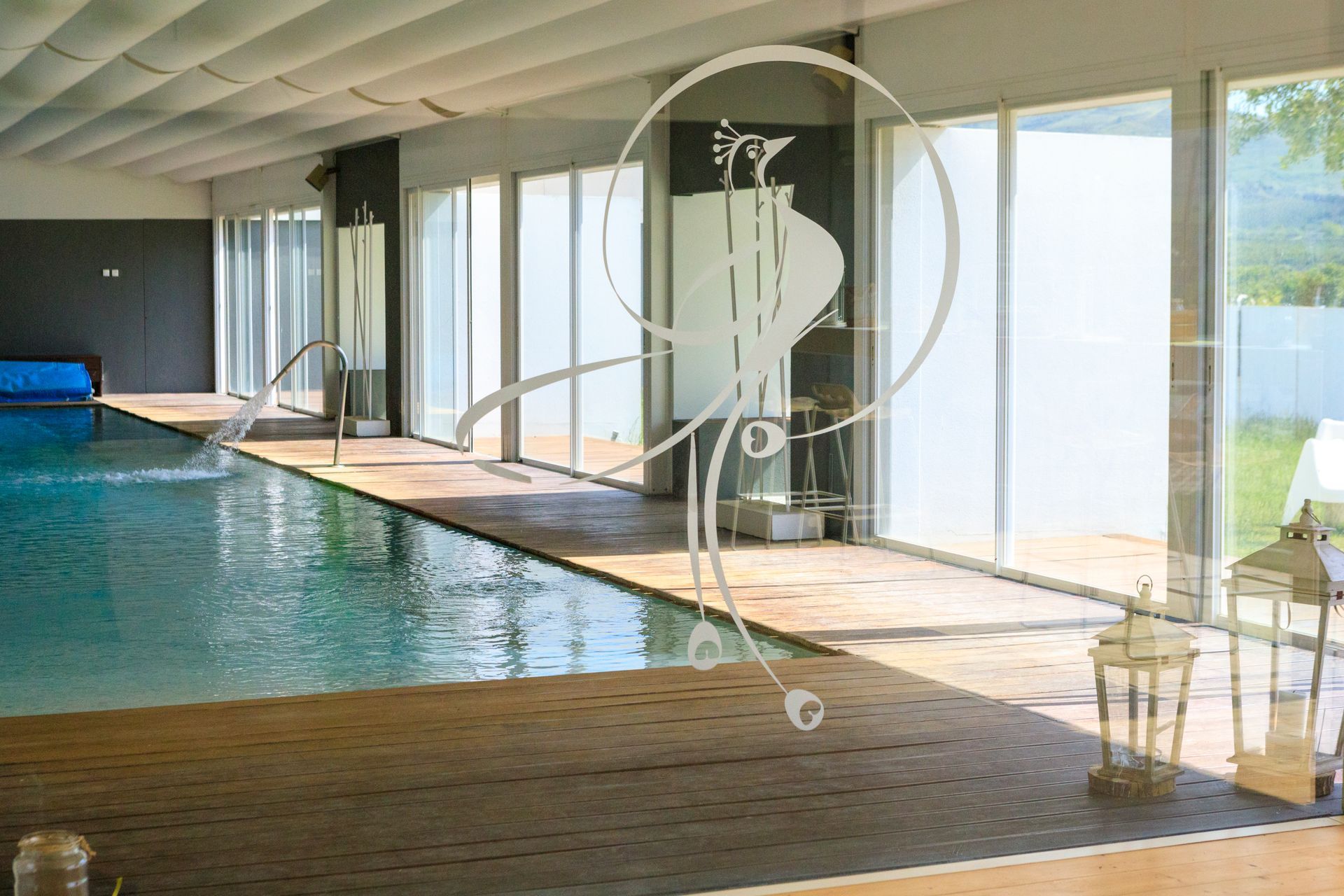 Piscina coperta con ampie vetrate, terrazza in legno e lettini prendisole con vista sull'acqua.