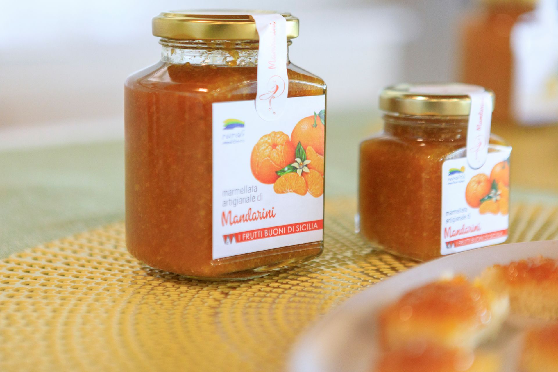 Due vasetti di marmellata d'arance con coperchi dorati su una tovaglietta intrecciata, uno più grande davanti.