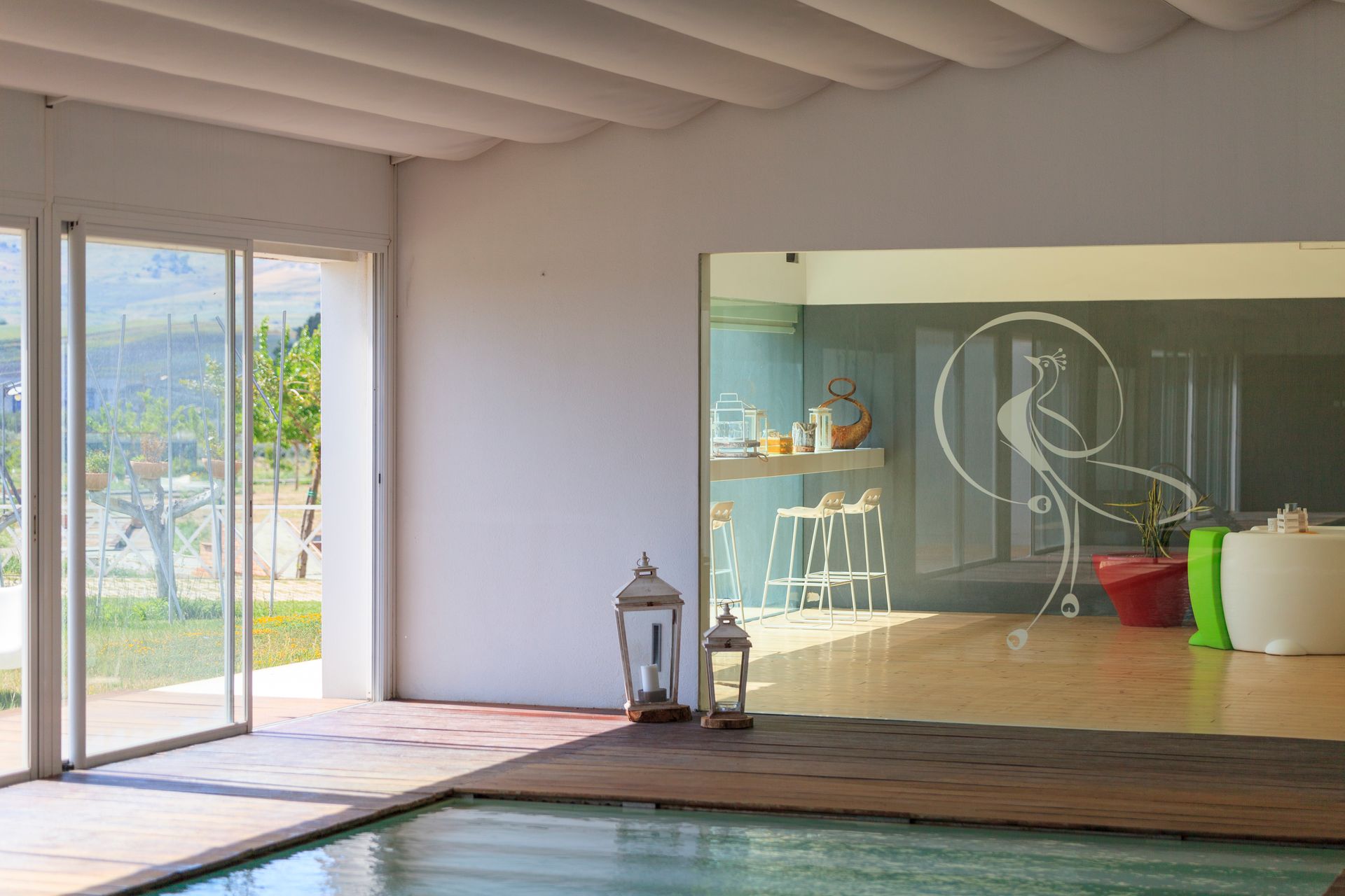 Piscina interna moderna con pavimentazione in legno, lanterne e porte finestre che si aprono su una luminosa stanza giardino.