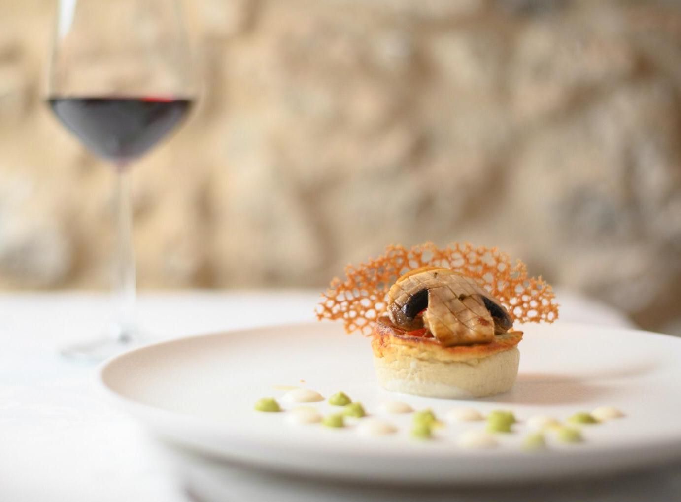 Un dessert gourmet su un piatto bianco con un calice di vino sullo sfondo, contro un muro di pietra sfocato.
