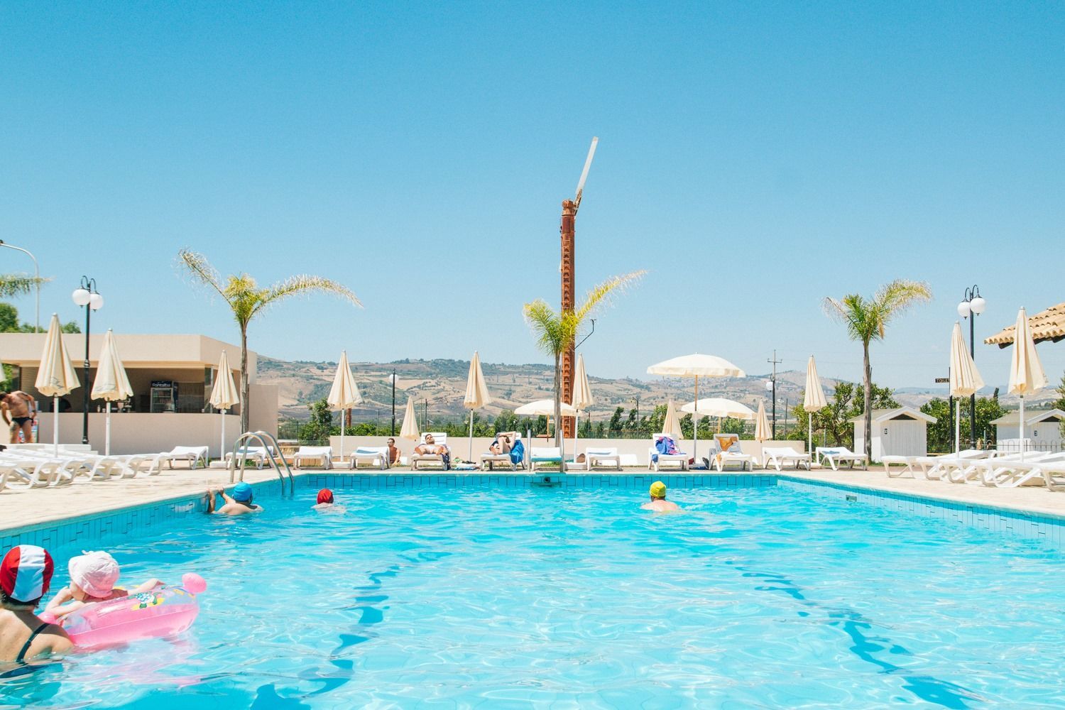 Luminosa piscina del resort con nuotatori, lettini bianchi, palme e il mare sullo sfondo, sotto un cielo azzurro limpido.