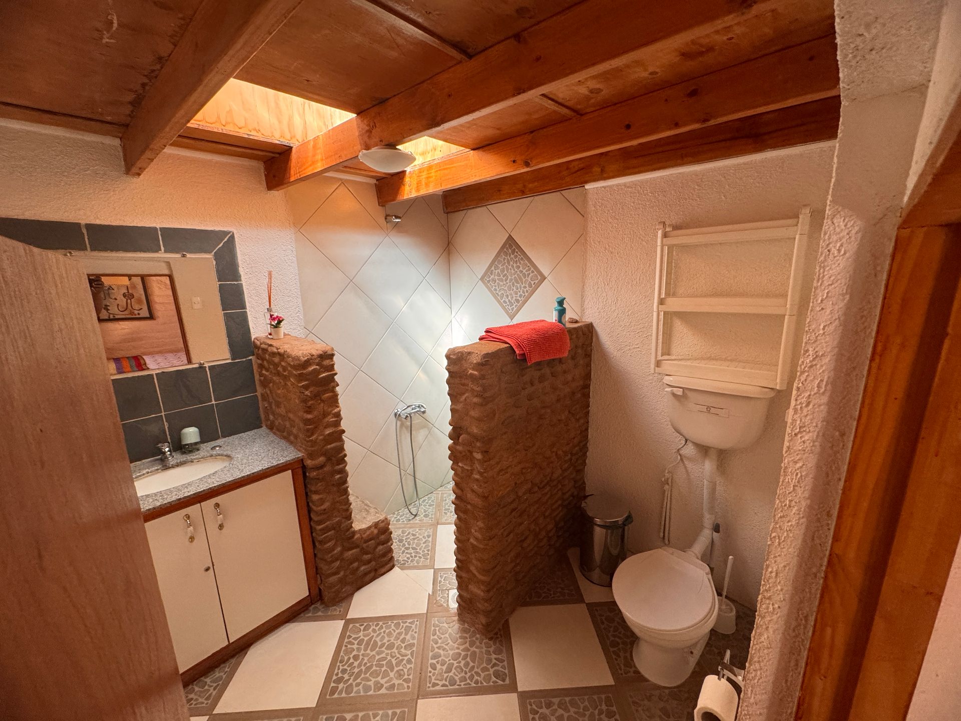 Pequeño baño rústico con inodoro blanco, lavabo, suelo de baldosas, techo de madera y mampara de ducha de ladrillo.