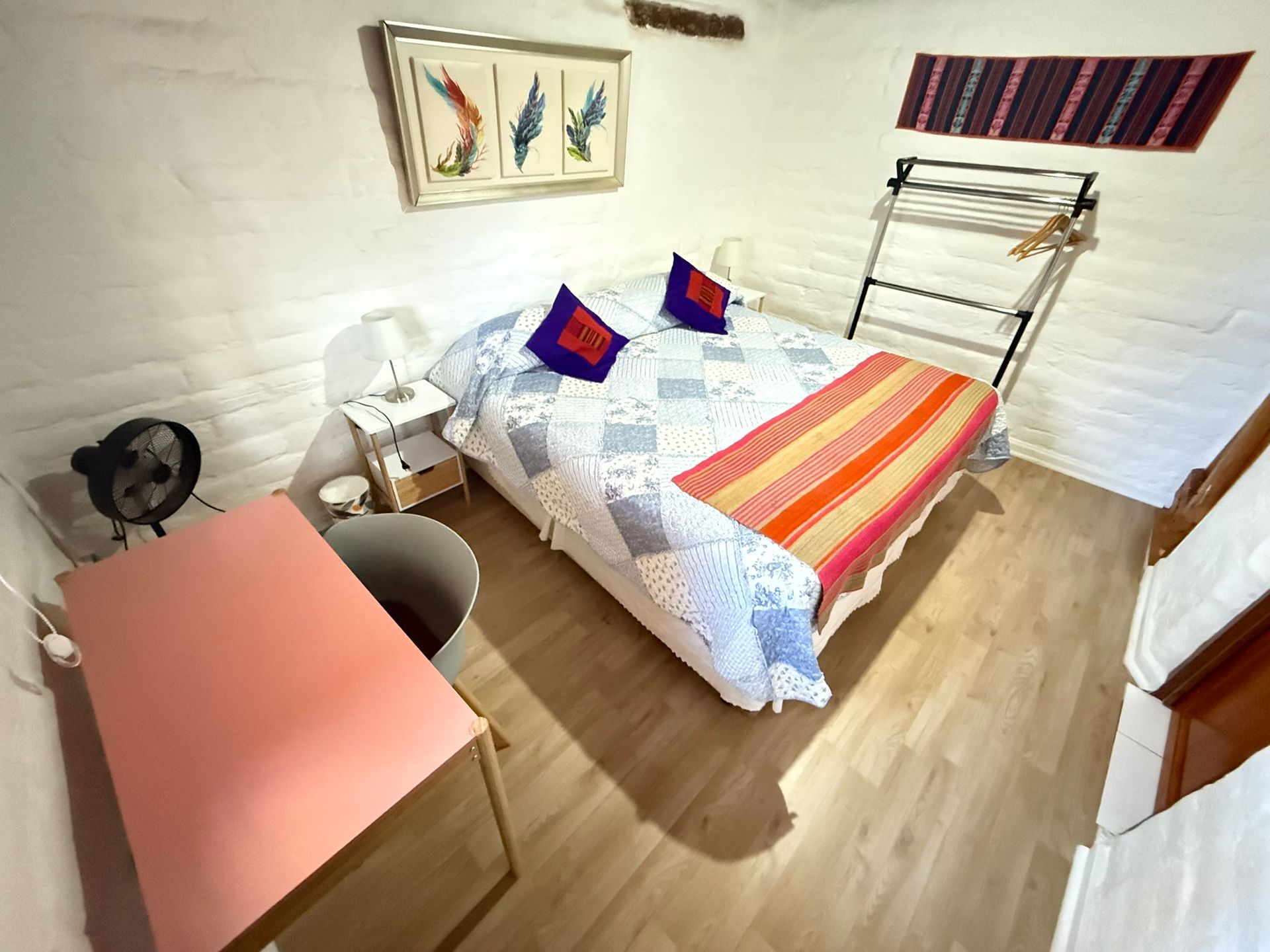 Habitación pequeña con cama, escritorio, silla, ventilador y cuadros en la pared sobre suelo de madera.