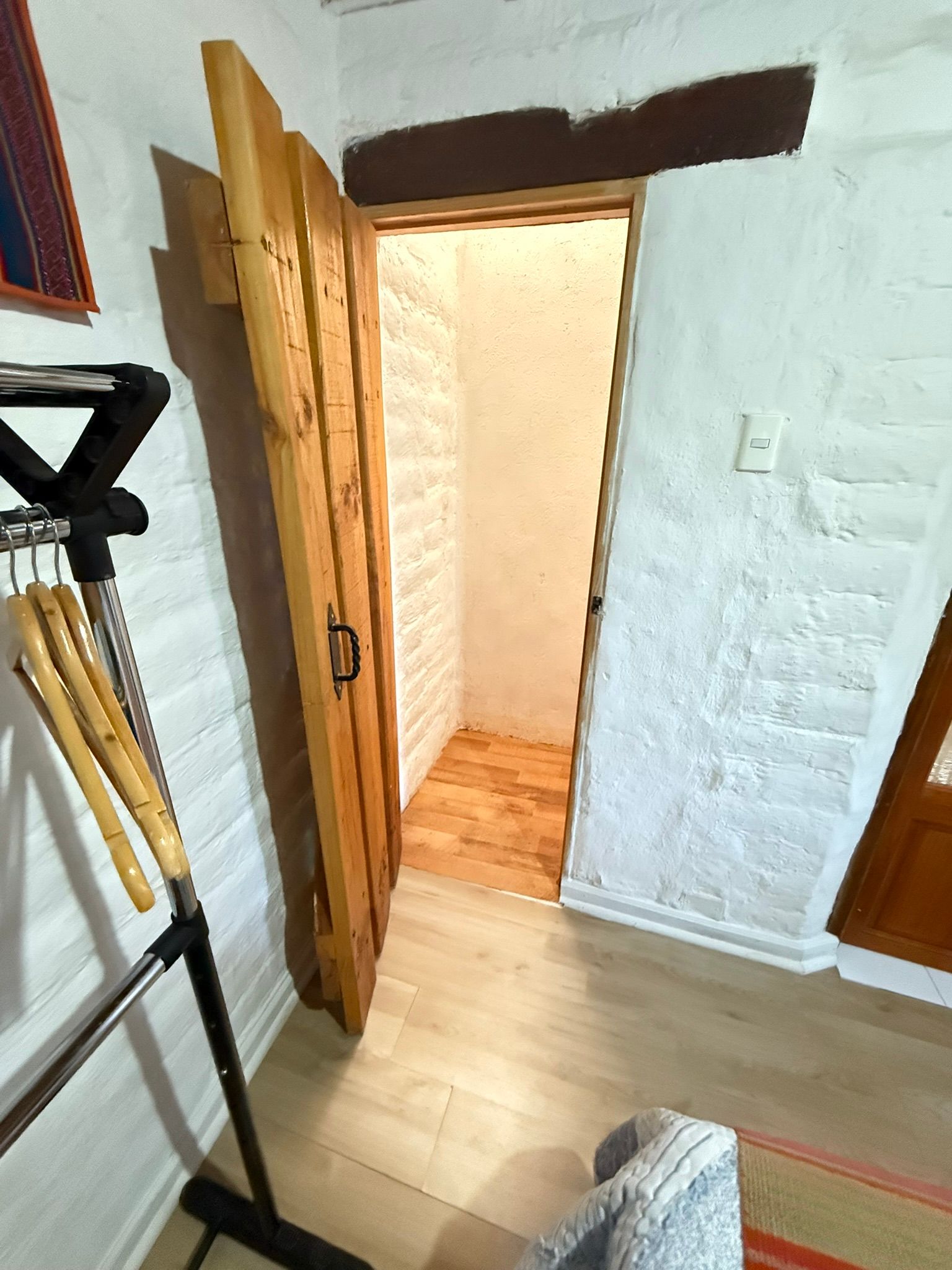 Se abre una puerta de madera que da a un pequeño y luminoso pasillo con suelo de madera clara y paredes de ladrillo blanco.