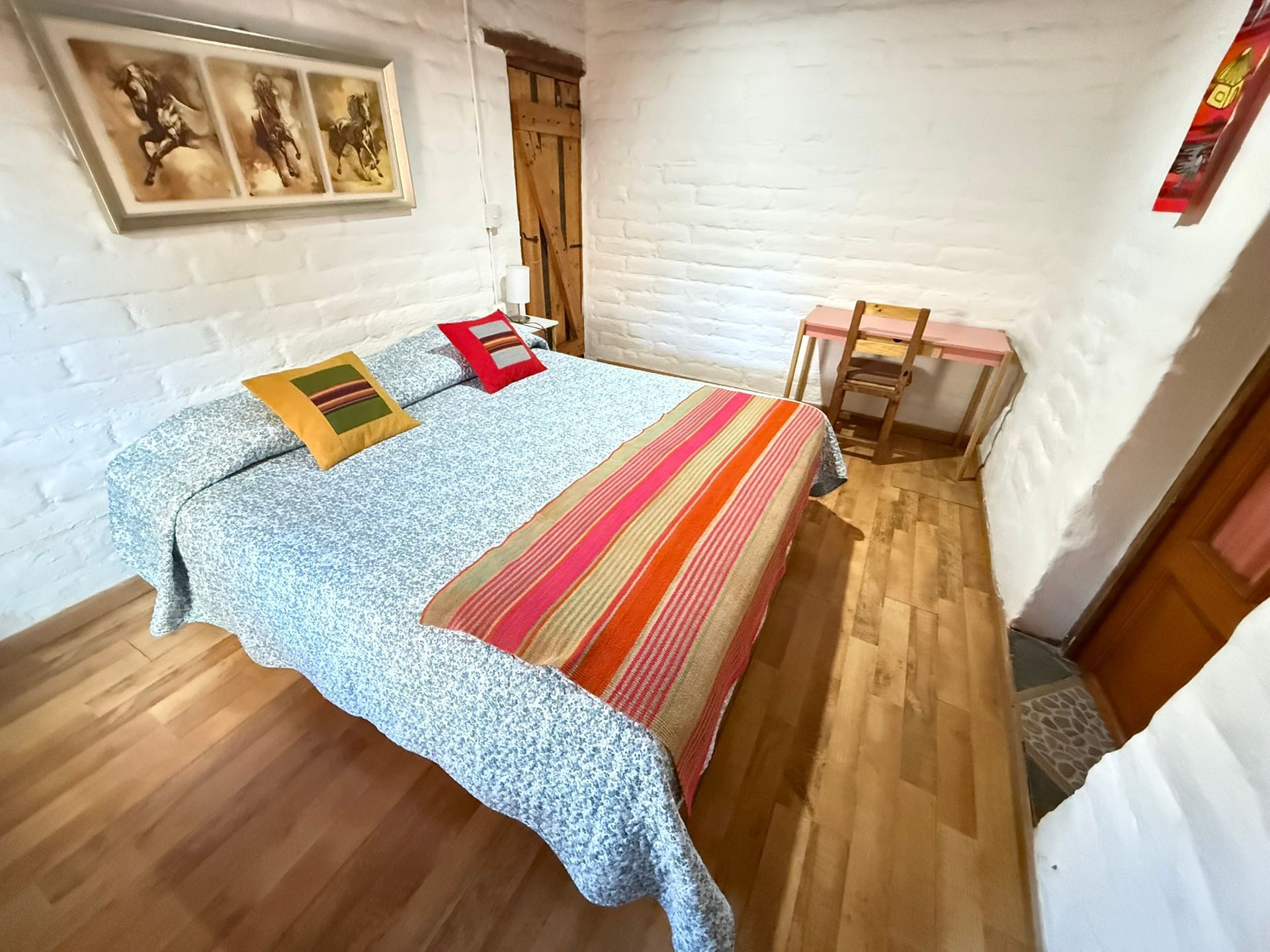 Habitación acogedora con cama, mantas coloridas, suelo de madera y un pequeño escritorio junto a la ventana.