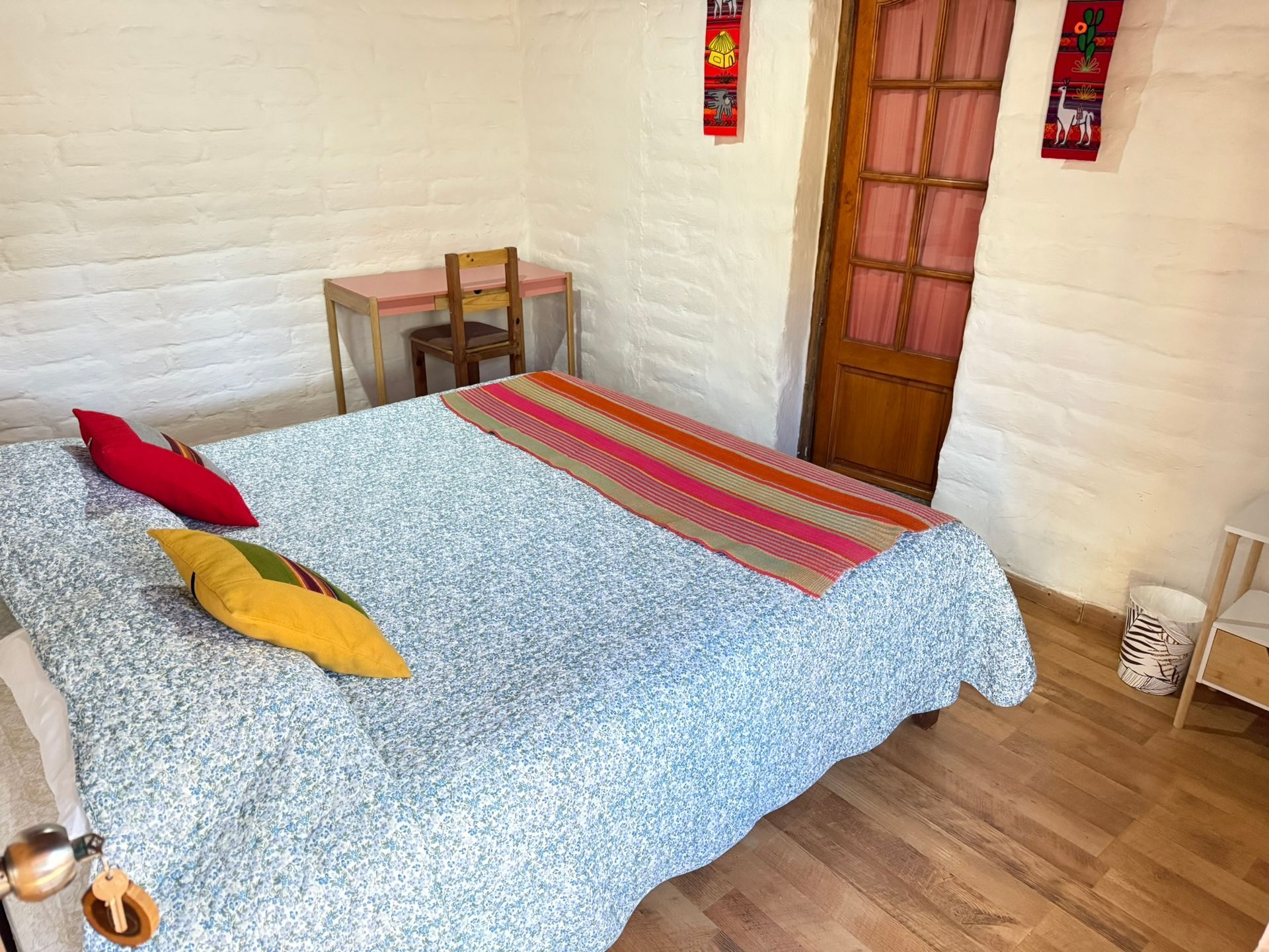 Habitación acogedora con cama, manta estampada, almohadas coloridas, escritorio pequeño y suelo de madera.