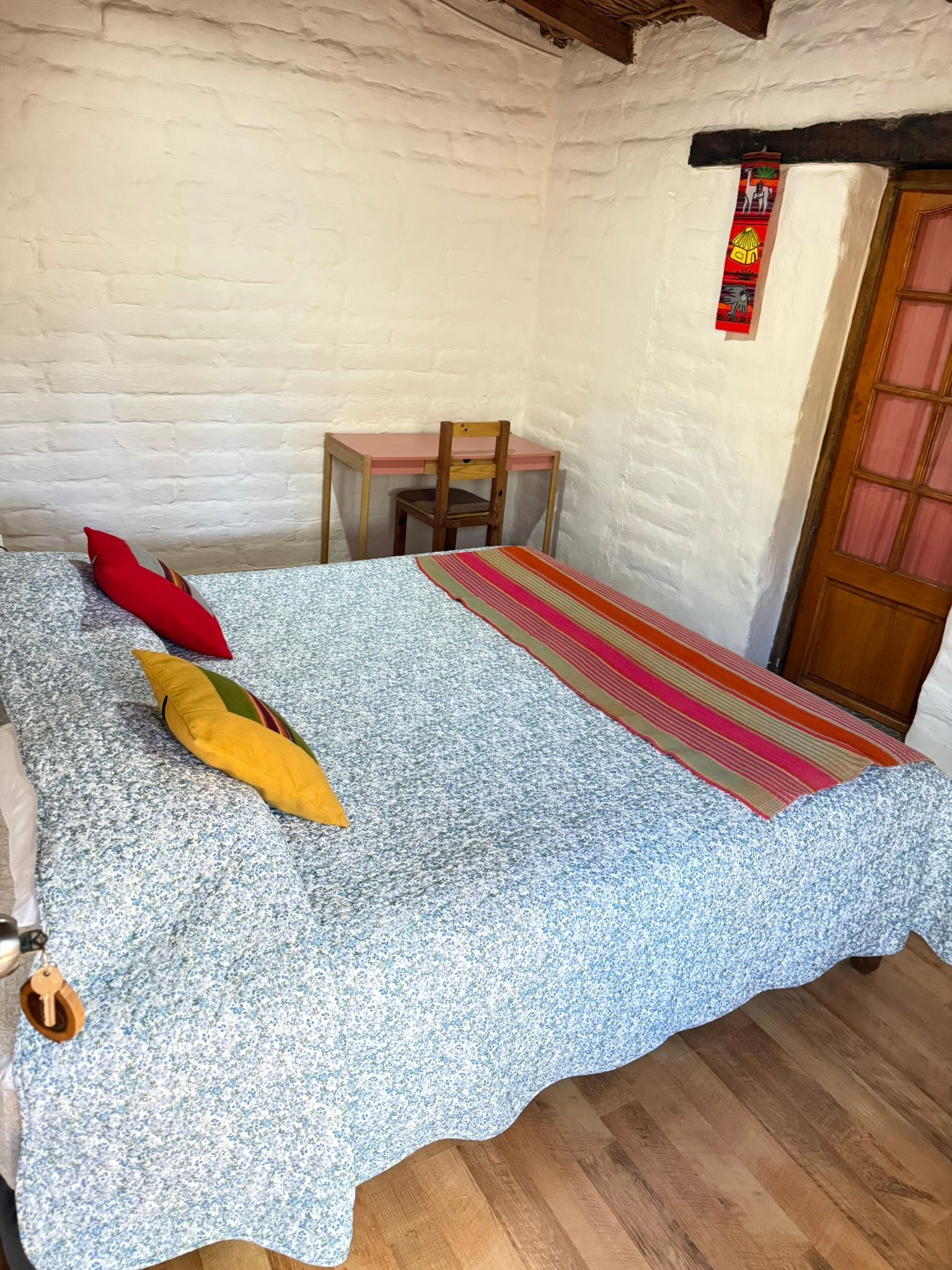 Habitación pequeña con una cama, almohadas de colores, una silla de madera y paredes de ladrillo blanco.