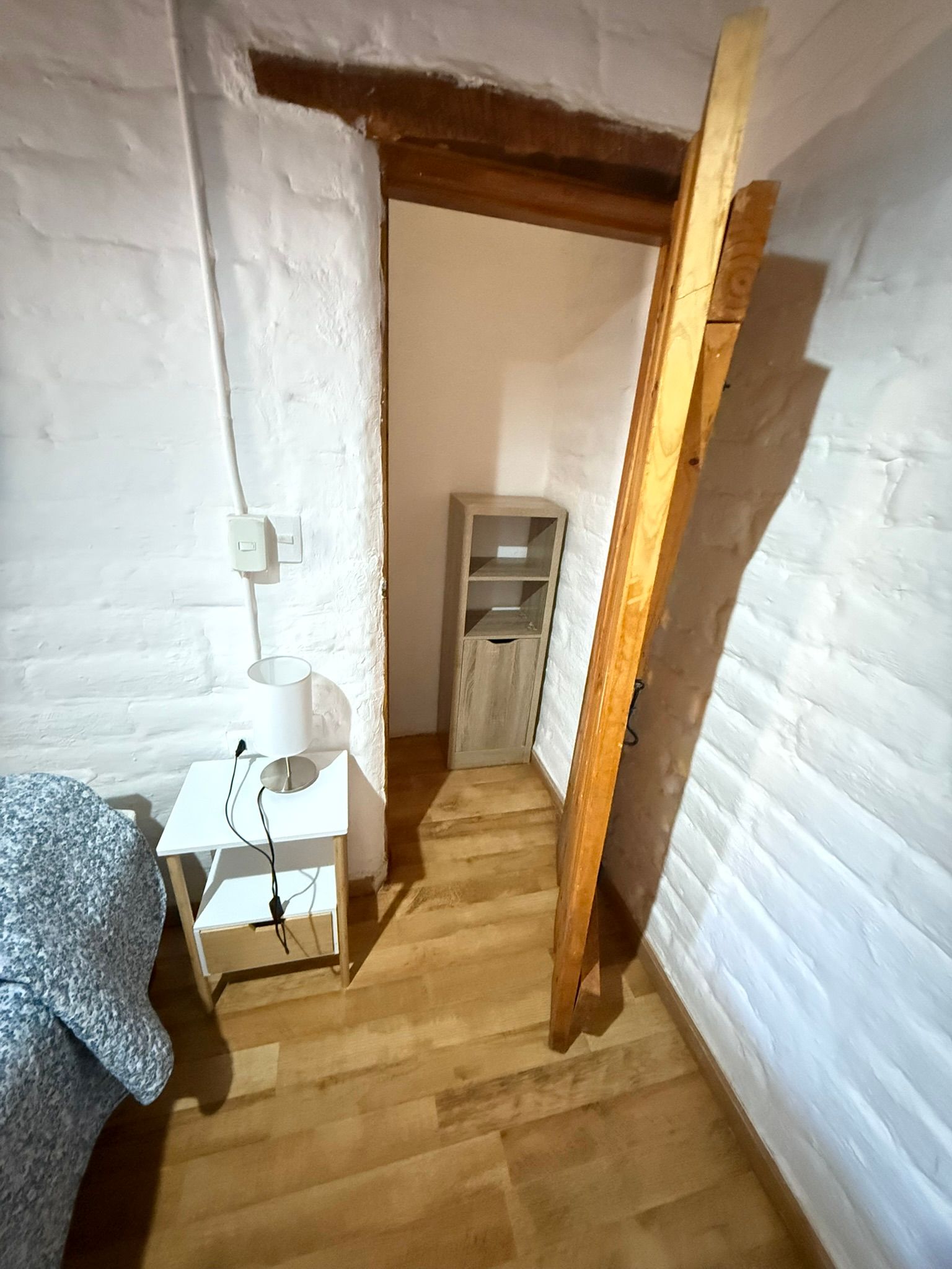 Pasillo estrecho con paredes de ladrillo blanco, suelo de madera, puerta abierta y una pequeña estantería al final.