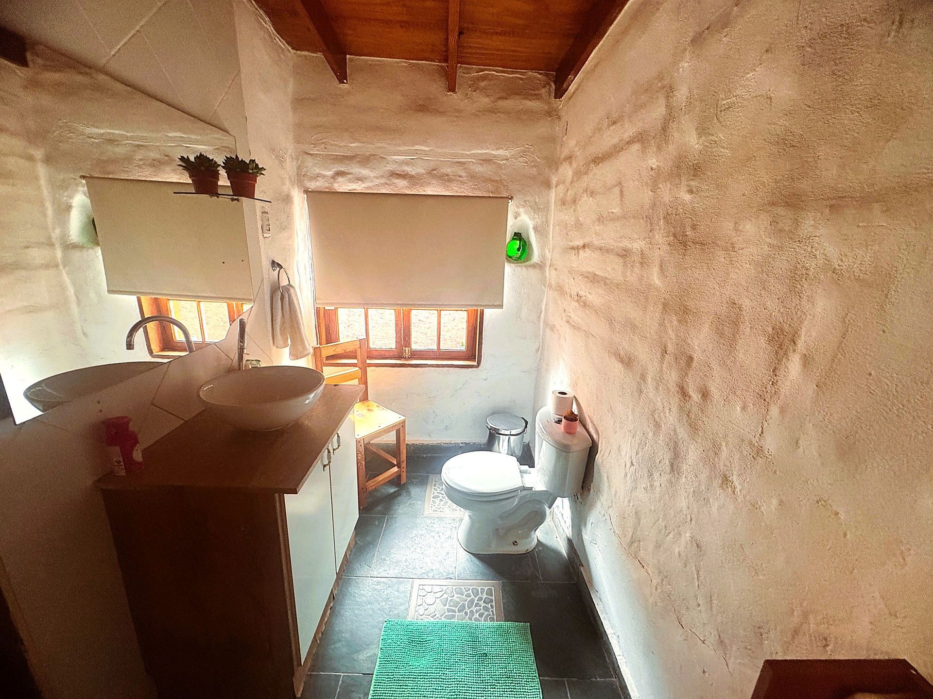 Pequeño baño rústico con lavabo, inodoro, bañera y alfombra verde en una habitación estrecha de color beige.