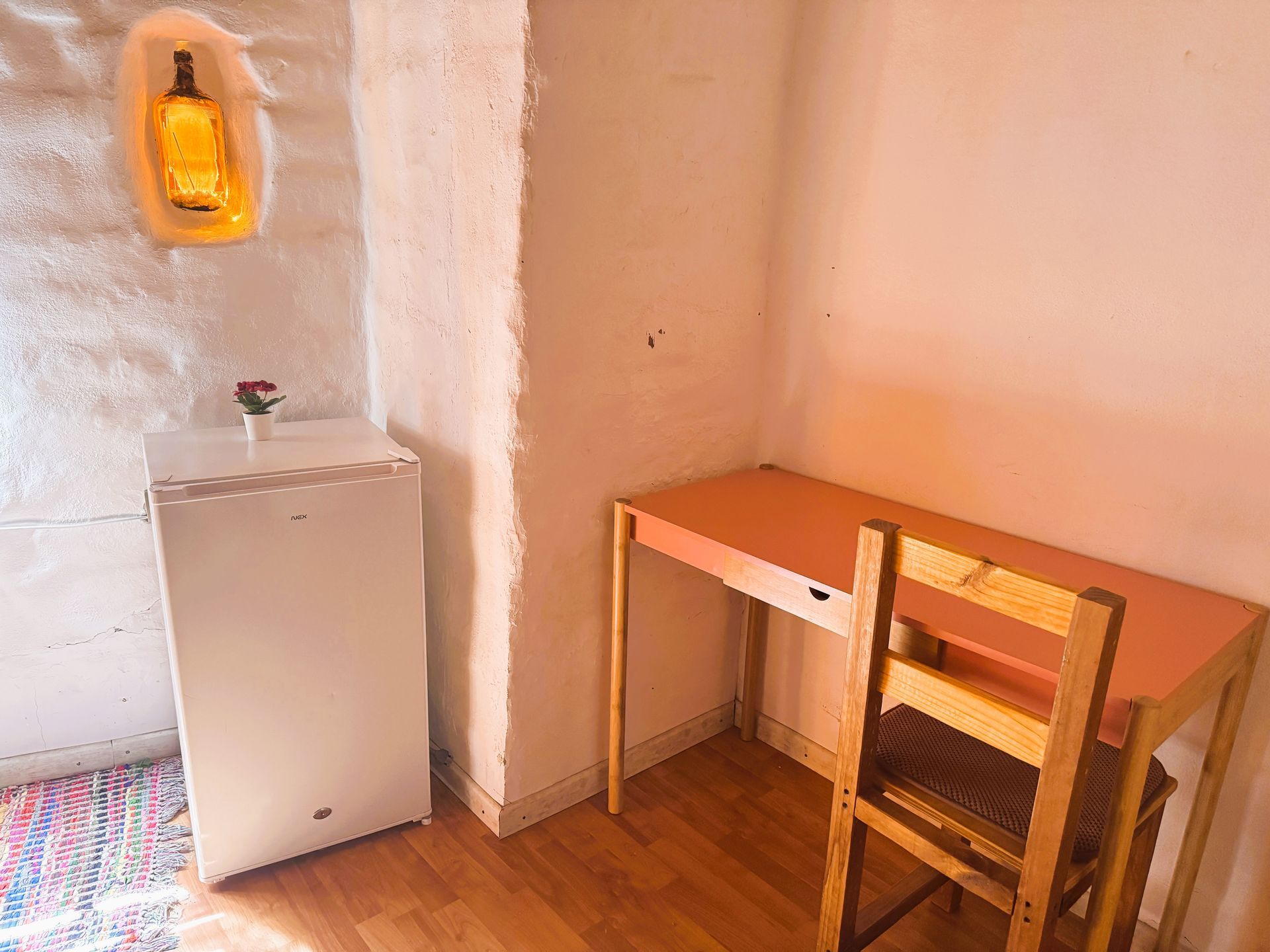 Habitación pequeña con un escritorio y una silla de madera junto a una mininevera y una lámpara de pared empotrada.