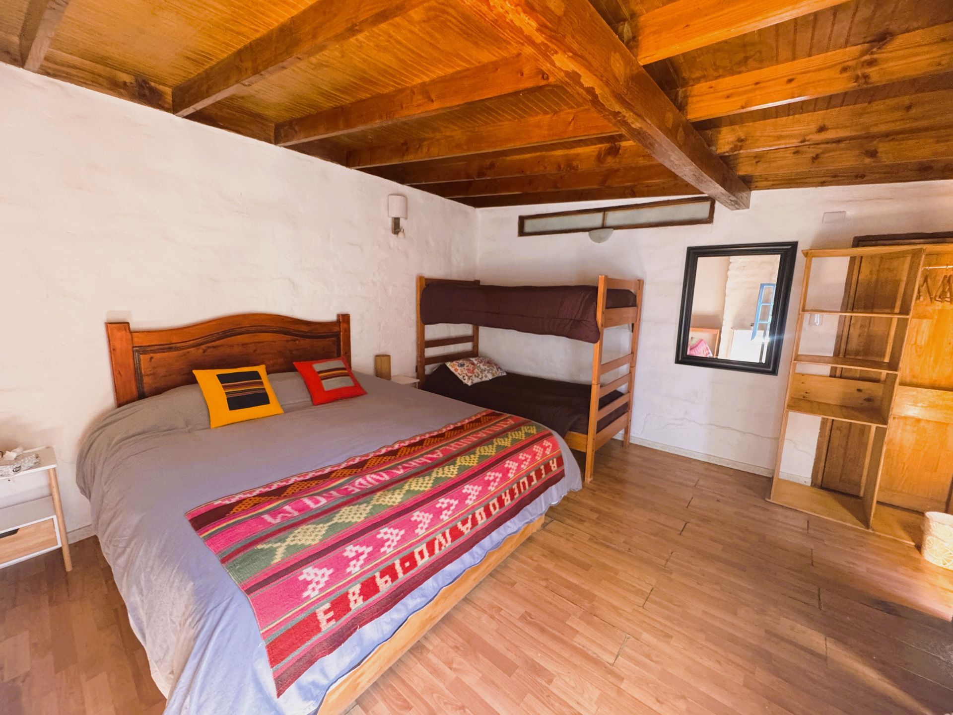 Habitación rústica con techo de madera, cama, litera y pequeña ventana en el suelo de madera.