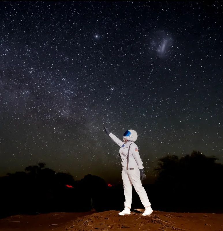 Astronauta con traje blanco señalando hacia arriba bajo un cielo nocturno estrellado sobre una superficie rocosa.
