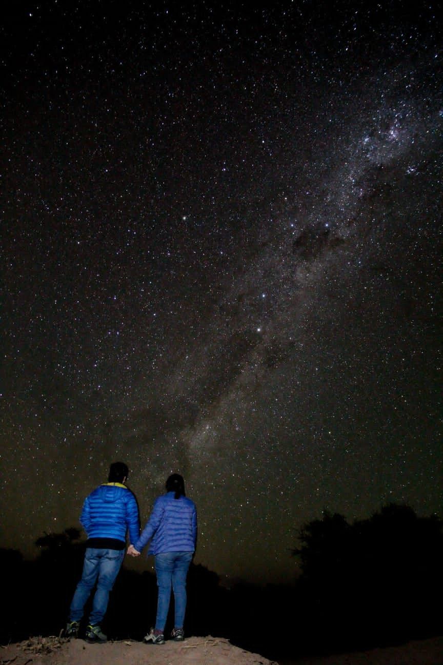 Dos personas con chaquetas azules permanecen de pie bajo un cielo nocturno estrellado, con la Vía Láctea de fondo.